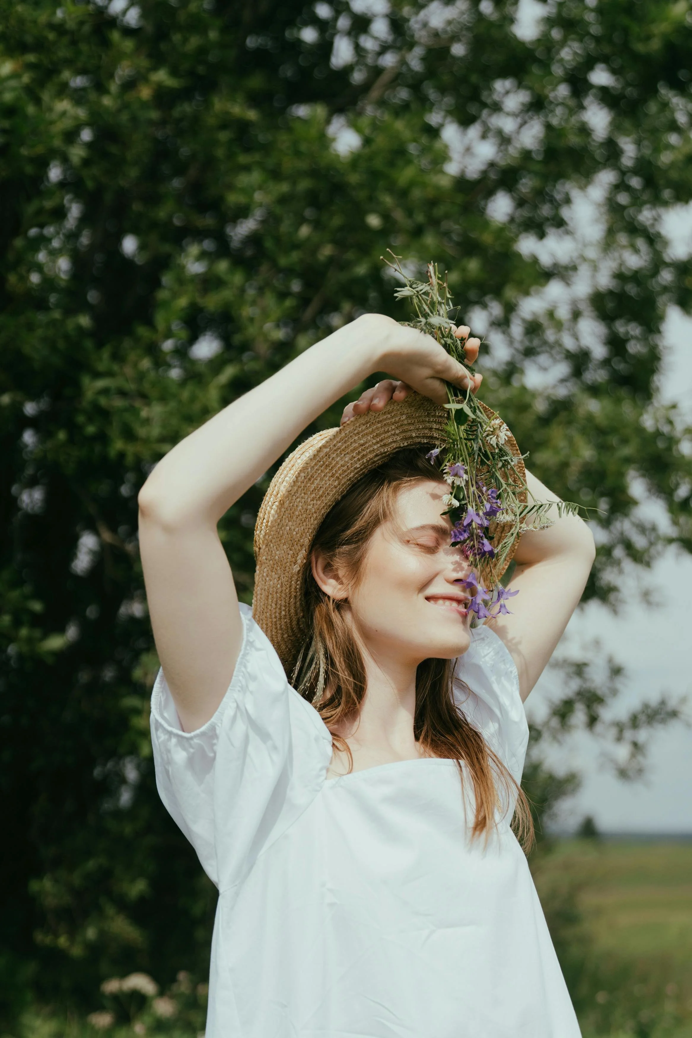 Een vrouw met lang, kastanjebruin haar die een witte jurk en een strooien hoed draagt, staat buiten in een natuurlijke omgeving met groene bomen op de achtergrond. Ze houdt bloemen boven haar hoofd en glimlacht met gesloten ogen.
