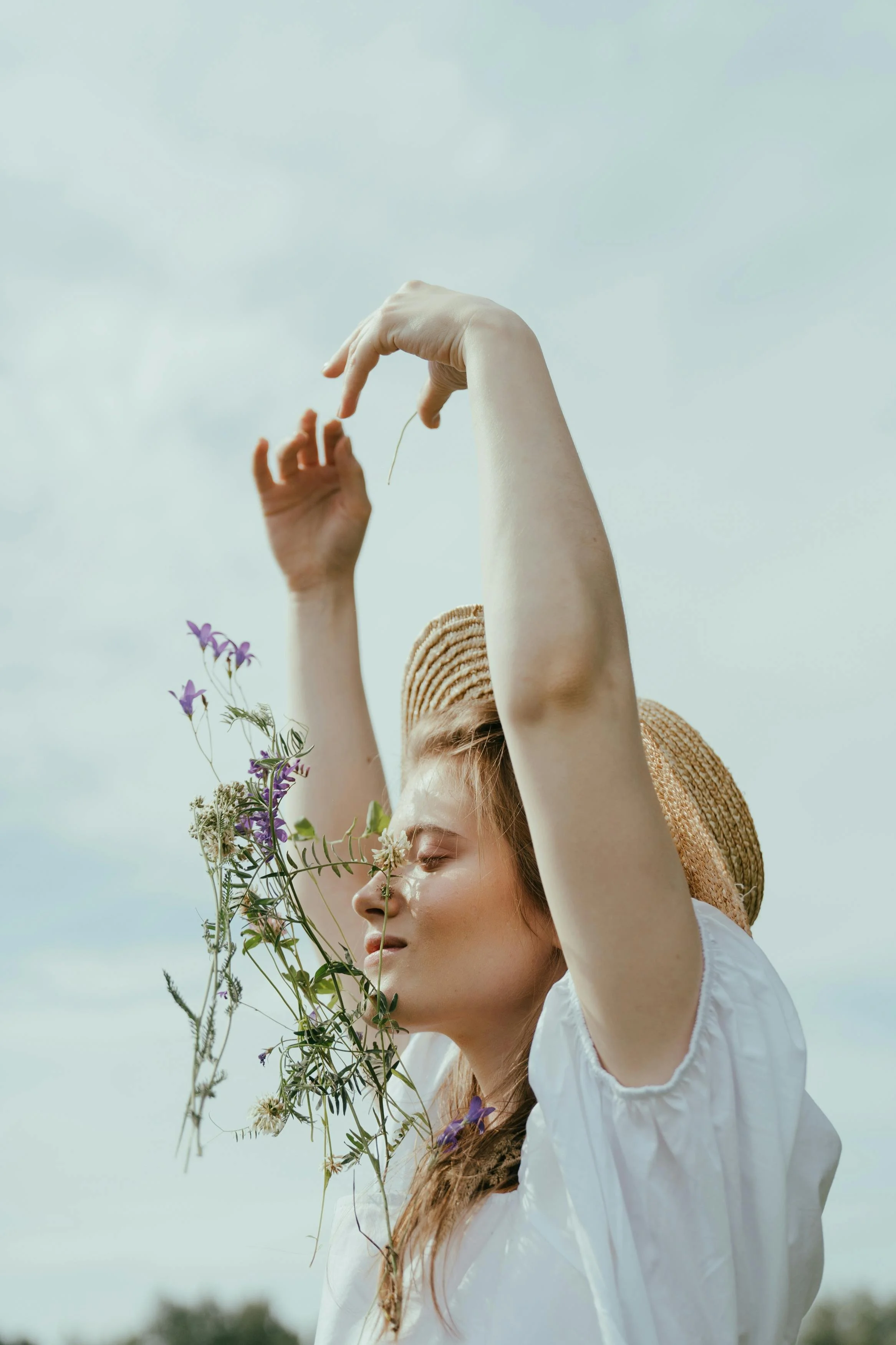 Jonge vrouw met lang haar, gesloten ogen, witte blouse, en een strohoed, die bloemen en planten los laat terwijl ze in de buitenlucht staat onder een bewolkte hemel.