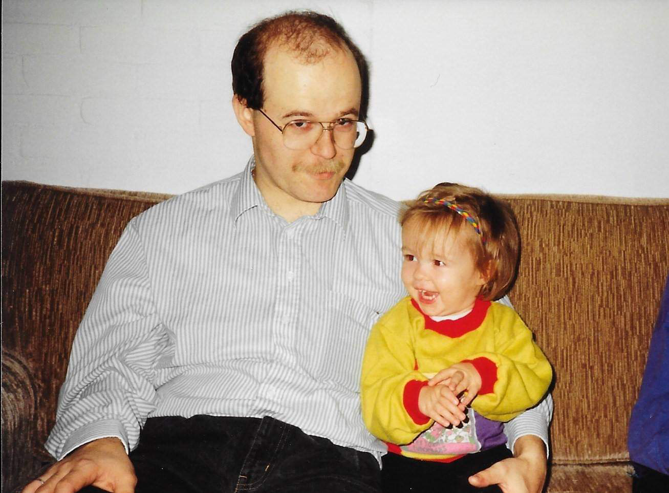 Een man met een bril en een lichte geruite overhemd zit op een bankje terwijl een jong meisje met een kleurrijk shirt en een hoofdband naast hem zit, beiden kijken naar elkaar.