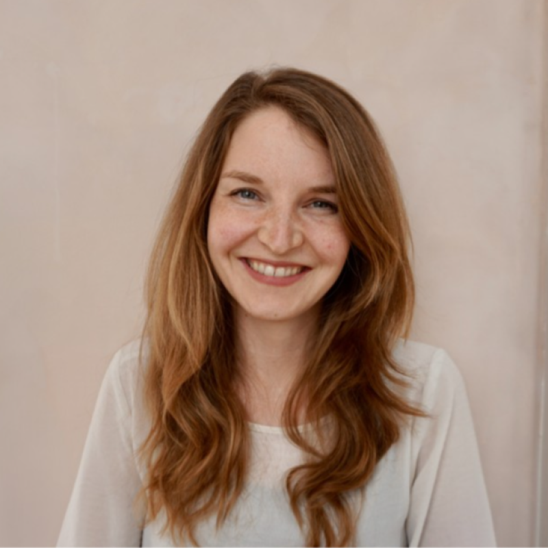 Vriendelijke jonge vrouw met rood haar die lacht en een witte blouse draagt, staand binnen bij een neutrale achtergrond.