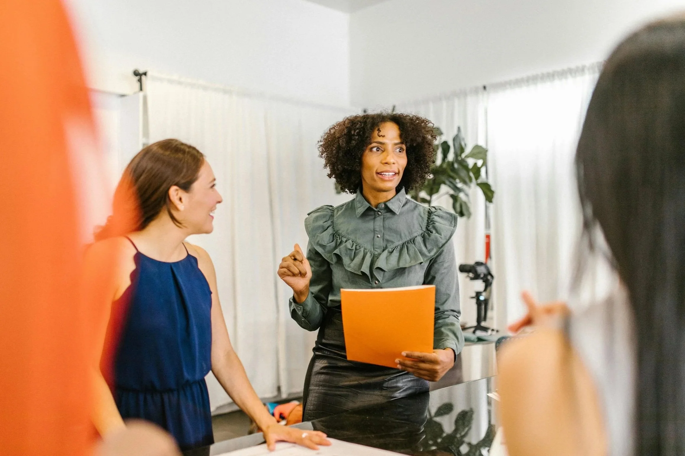 Leider staat in een kamer met witte muren en grote ramen, haar handen gebruikend terwijl ze praat met een groep mensen.