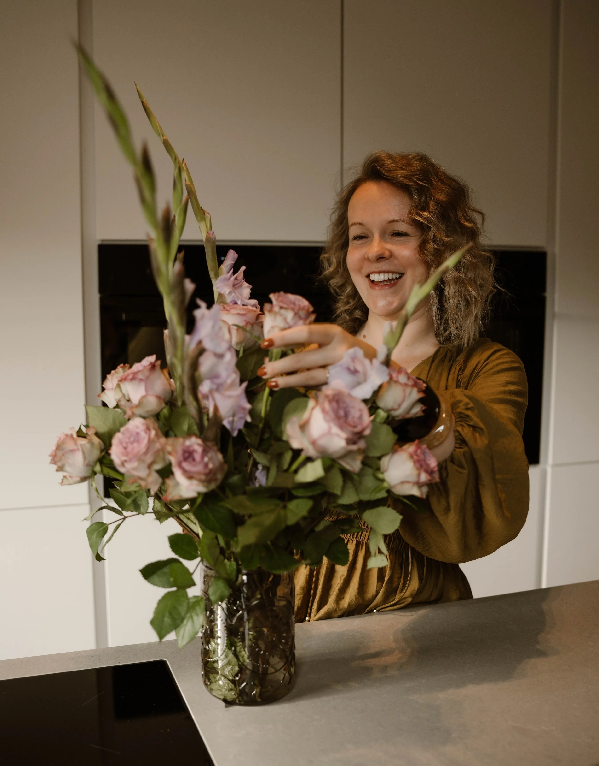 Een lachende vrouw met kastanjebruine, gekrulde haar houdt een grote boeket roze en witte bloemen.”}