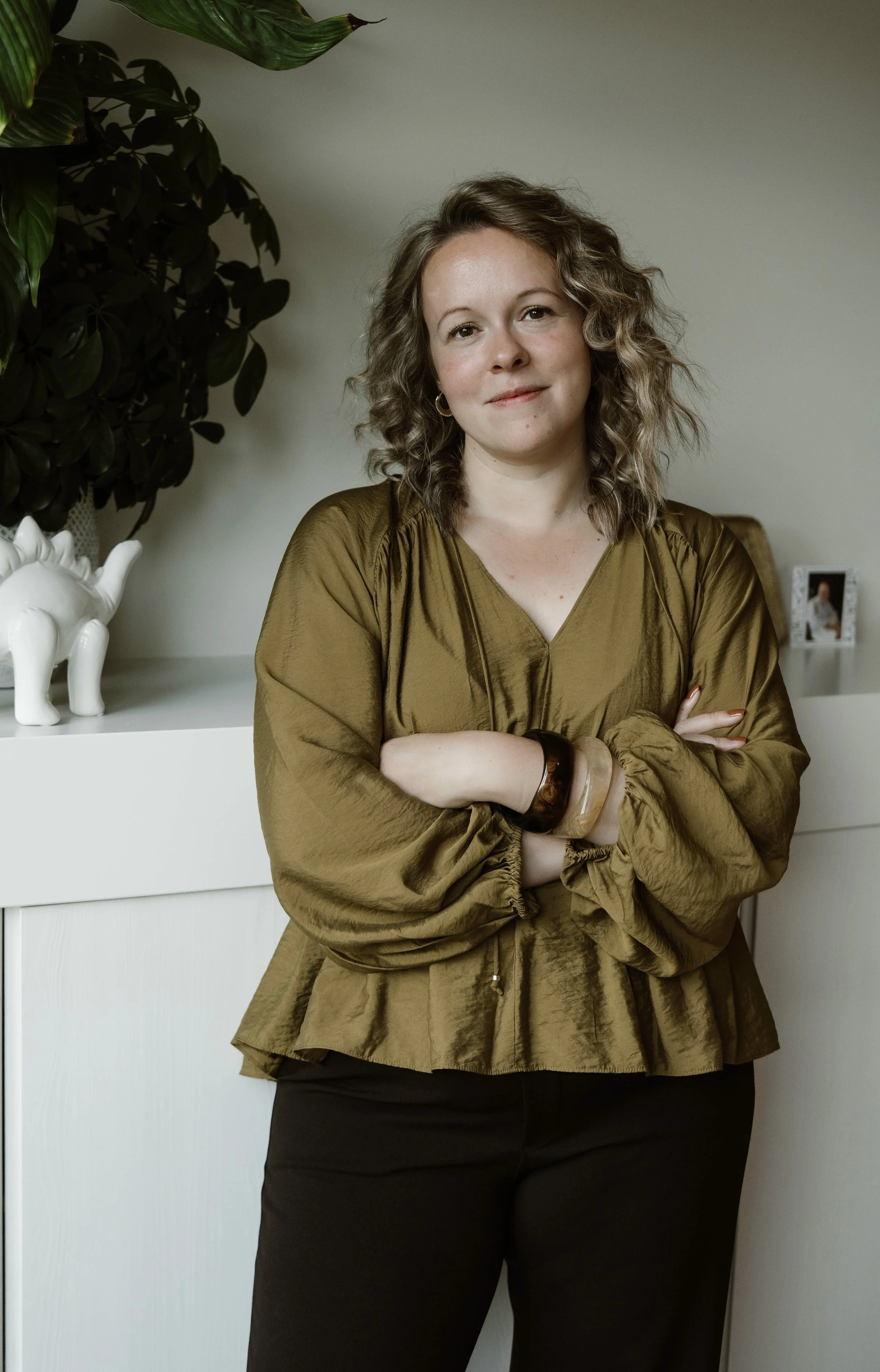 Vrouw met wavy haar staand binnen, gekleed in een goudenbruine blouse en zwarte broek, met haar armen gekruist, op de achtergrond een wit kastje met foto en decoraties, en een groene plant.