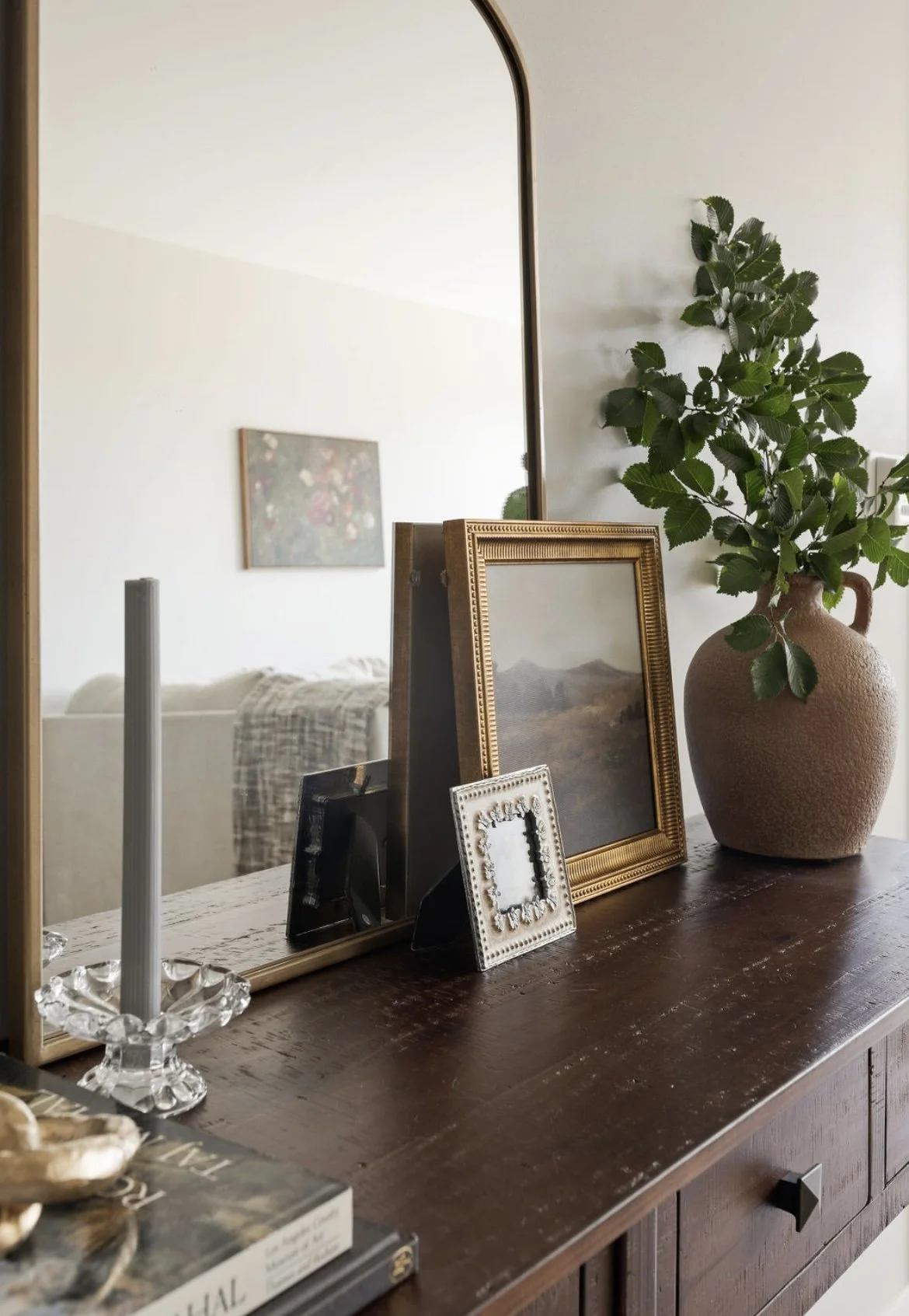 A wooden dresser with a large mirror, decorated with framed photographs, a landscape painting, a potted plant with green leaves, and a candle holder.