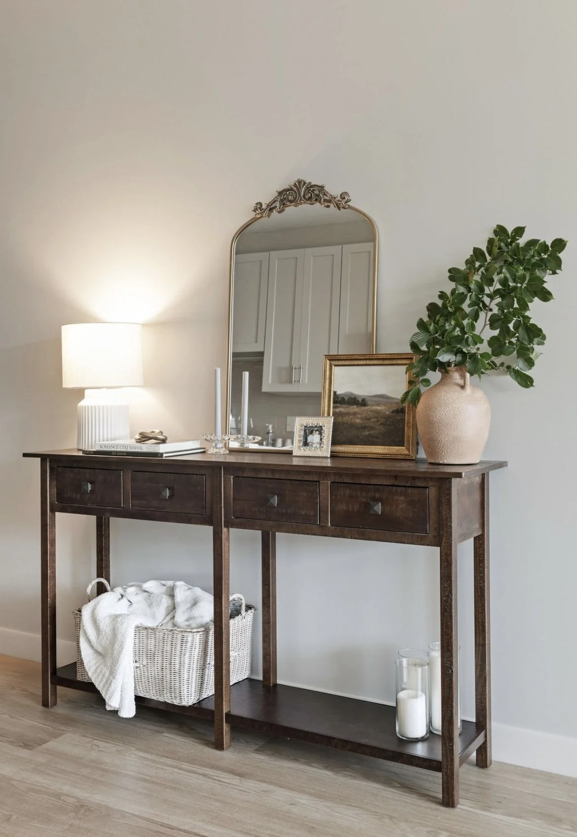 A wooden console table with a lamp, candles, picture frames, a potted plant, and a mirror on the wall above it.