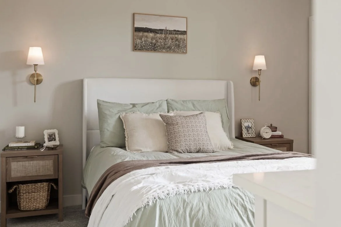 A cozy bedroom with a white upholstered bed, multiple pillows, and a brown throw blanket. There are two wooden nightstands with lamps, books, and picture frames, and a framed landscape photograph hanging above the bed.
