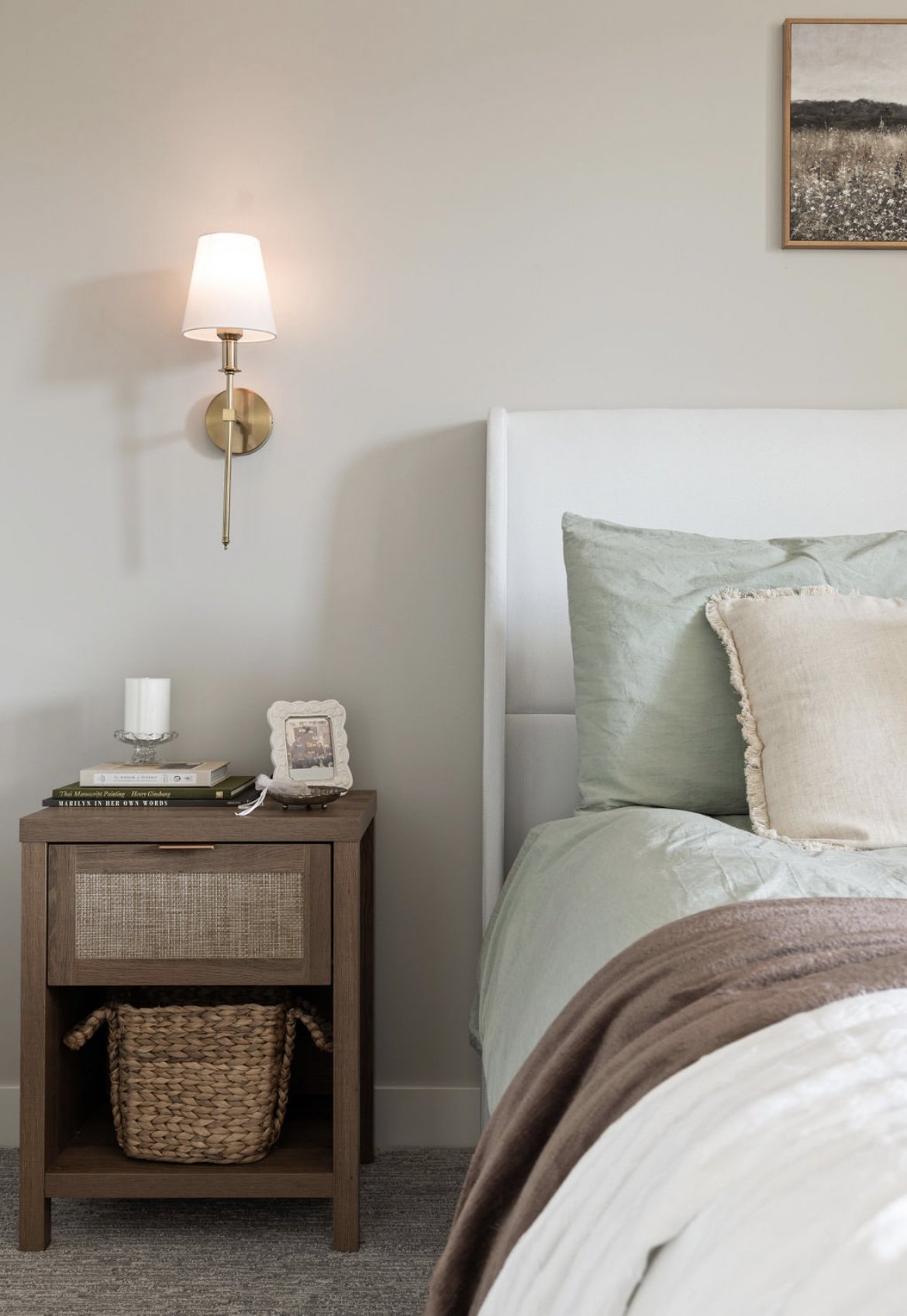 A bedroom corner with a white bed, green and beige pillows, a wooden nightstand with a basket beneath, a candle, a framed photo, books, a wall-mounted lamp, and a partial framed artwork.