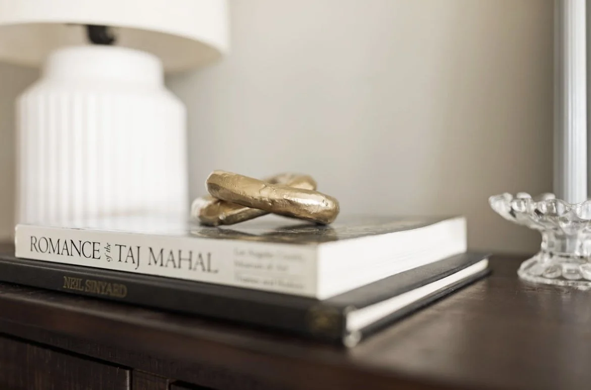 Decorative objects on a wooden surface, including a white lamp, a large book titled "Romance of the Taj Mahal," a smaller book underneath, and a clear glass dish on the right.
