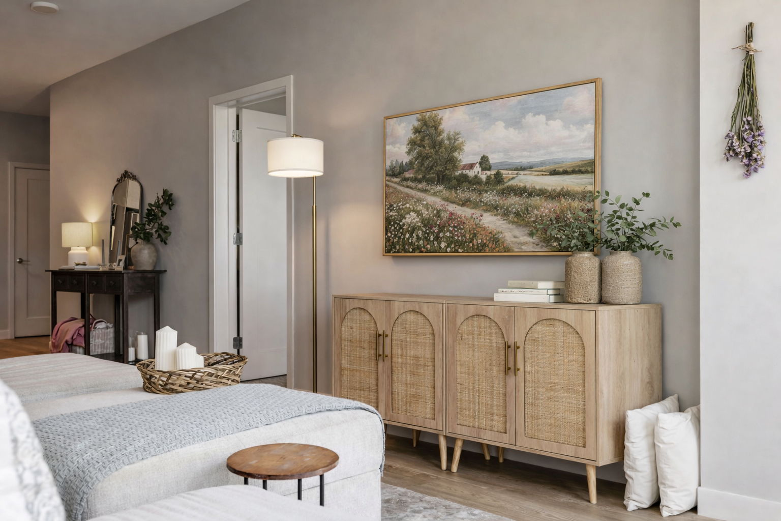 A living room with a wooden sideboard, a large landscape painting above it, decorative vases with greenery, a framed mirror, and a tall floor lamp, with couches and candles in the foreground.