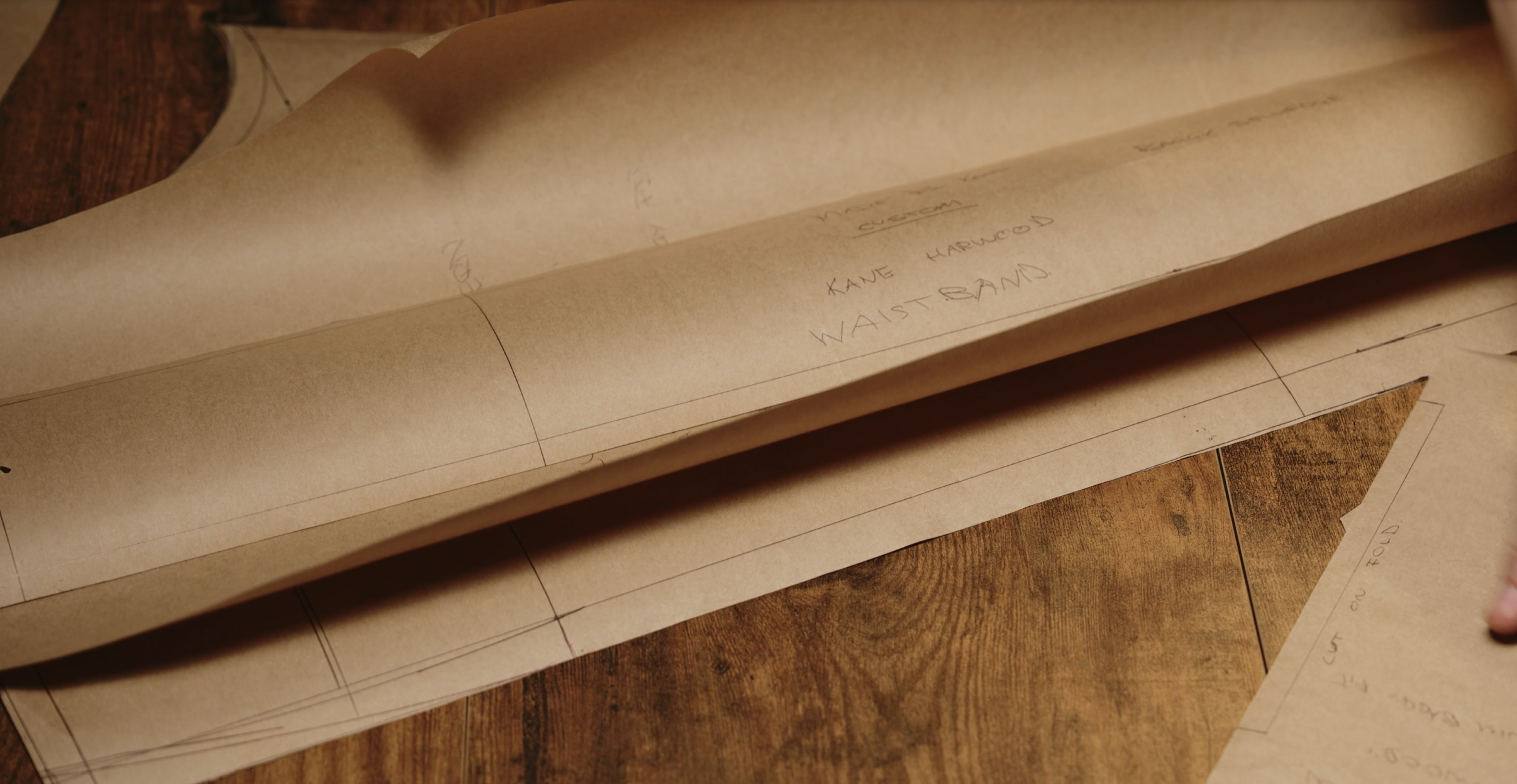 Close-up of pattern-making paper with wooden background, showing drafting lines and notes including 'KANE HARWOOD WAISTBAND'.