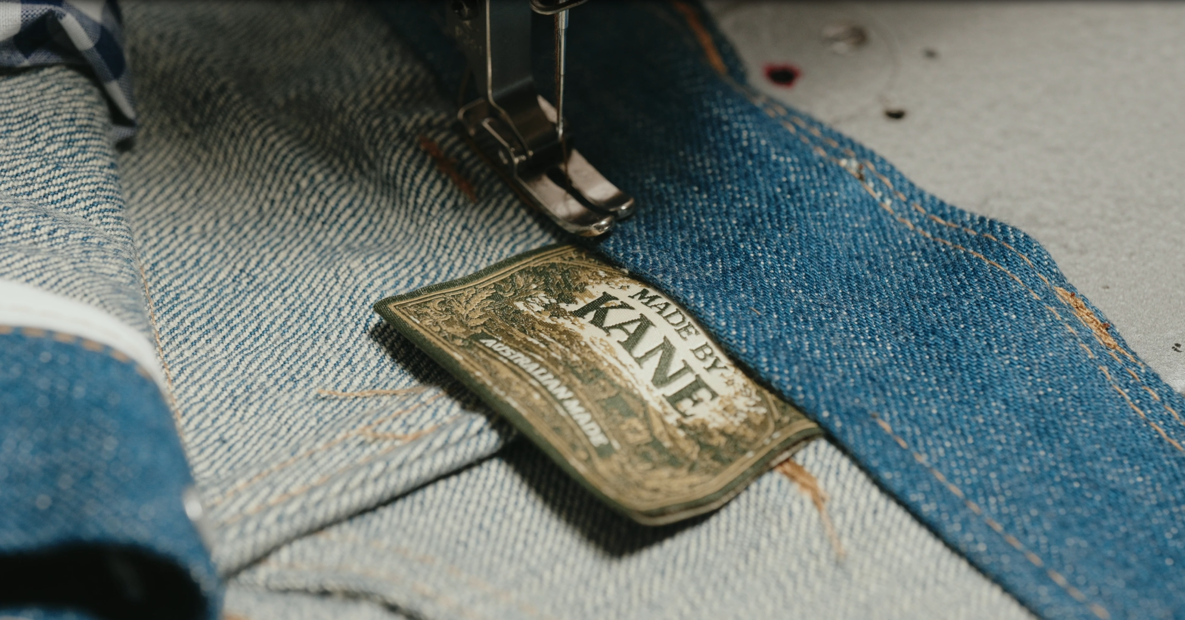 Close-up of denim fabric with a 'Made by KAVE' tag and sewing machine needle stitching the fabric.
