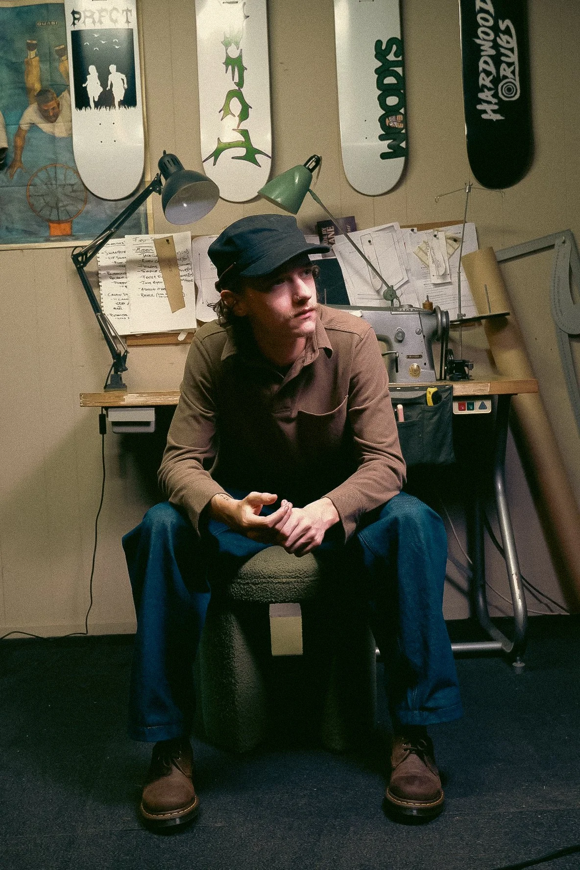 A young man sitting on a stool in a room with skateboard decks mounted on the wall behind him, a worktable with various tools and papers, and two adjustable lamps overhead.
