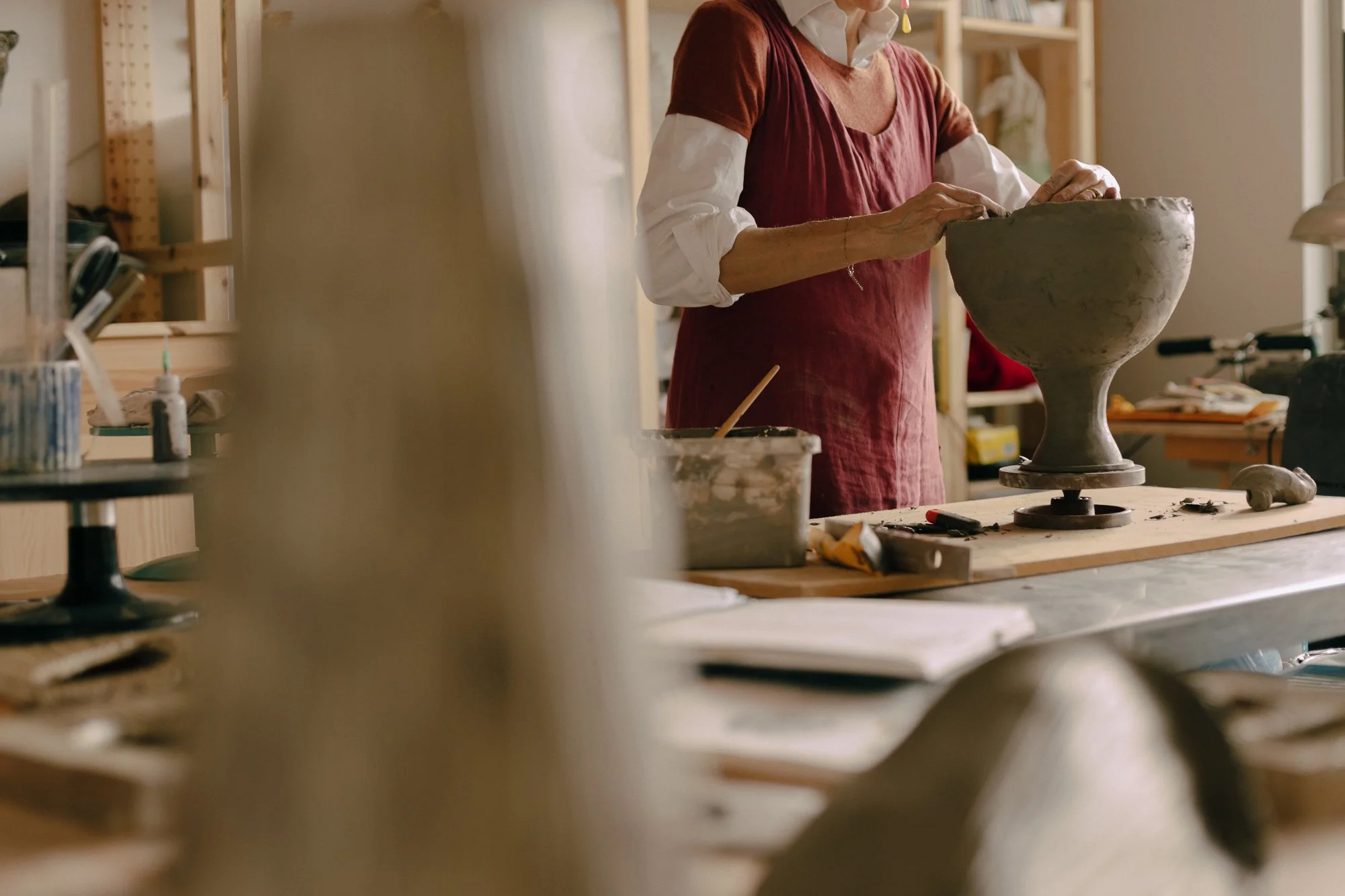 A person working on a pottery wheel in a ceramics studio, wearing a red smock over a white shirt.