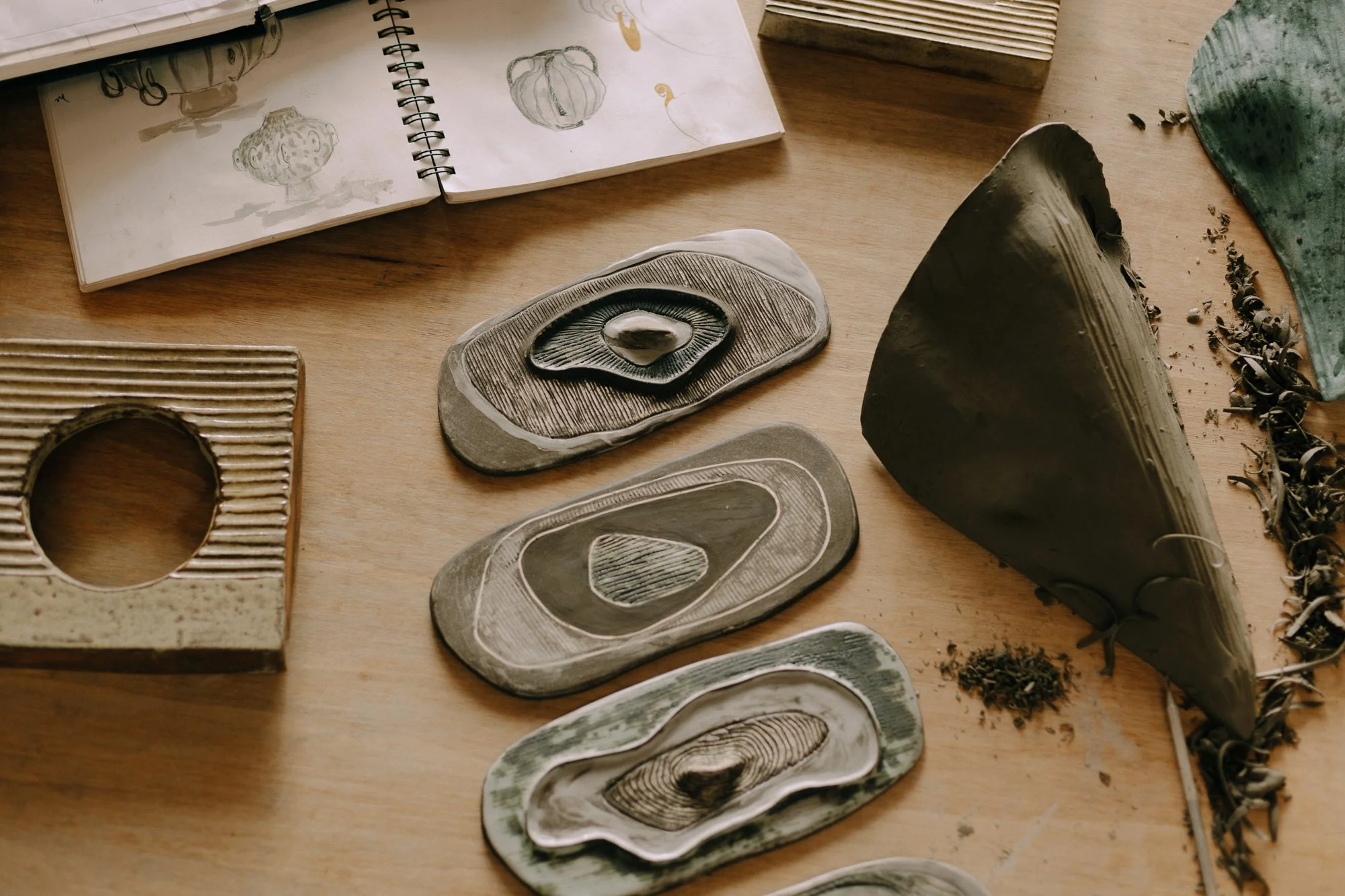 A working table displaying various ceramic sculptural pieces, an open sketchbook with drawings of vases art tools and an unfired lamp half worked..