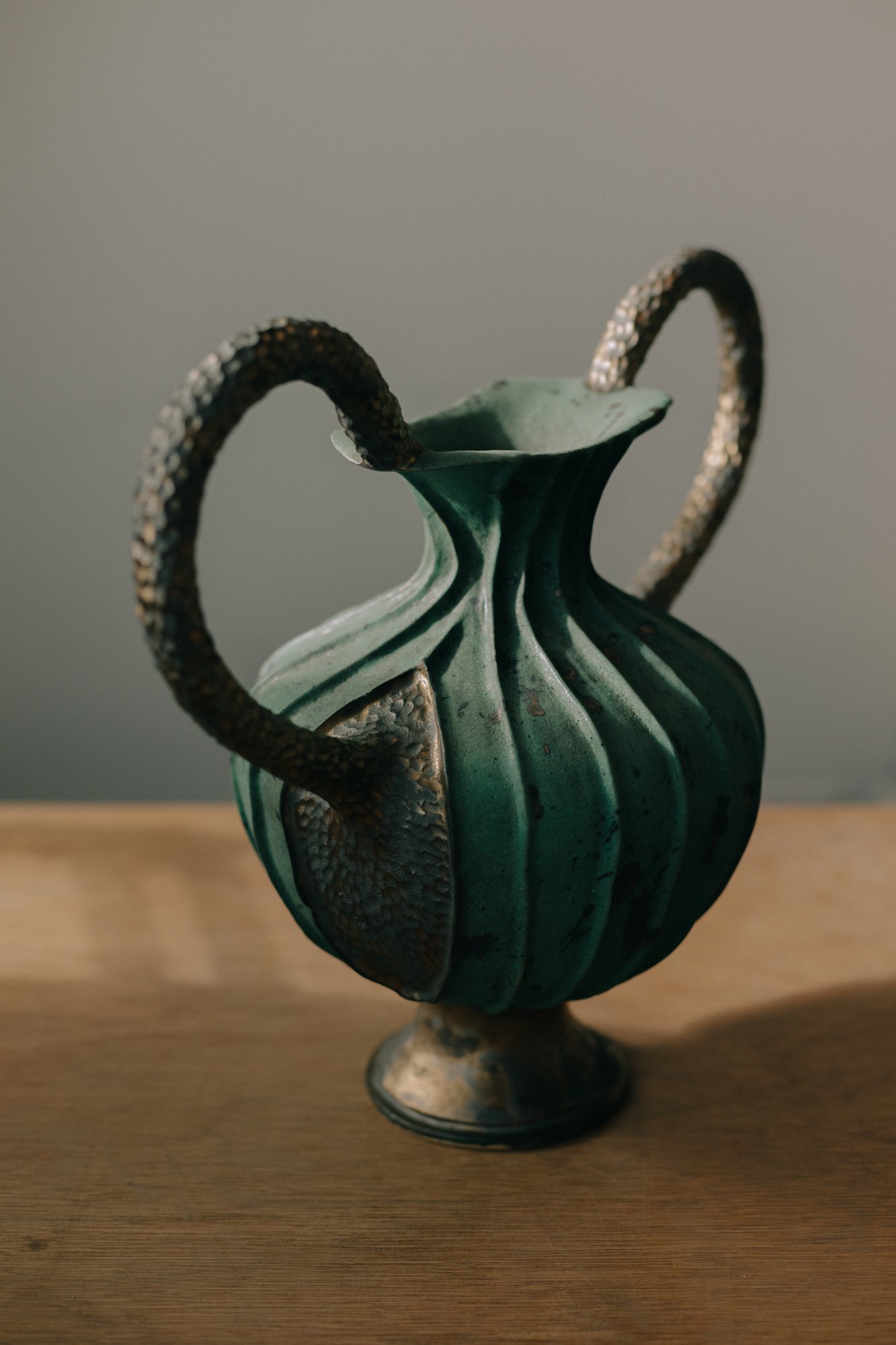 A large ceramic vase with a green, ribbed body, and two textured, arched handles, placed on a wooden surface.