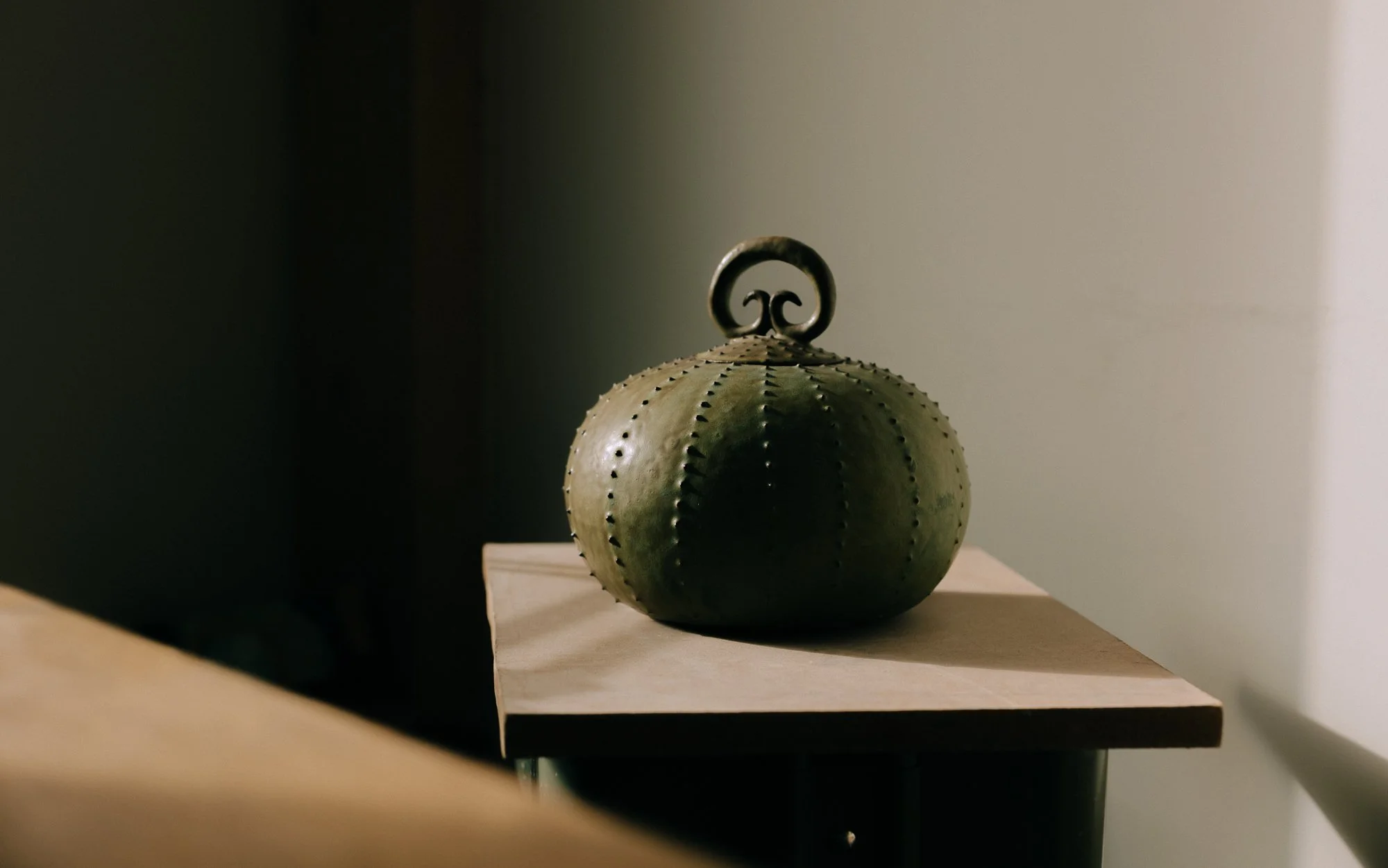 A decorative object shaped like a pumpkin, made of green material with small perforations and a curly handle on top, placed on a wooden surface.