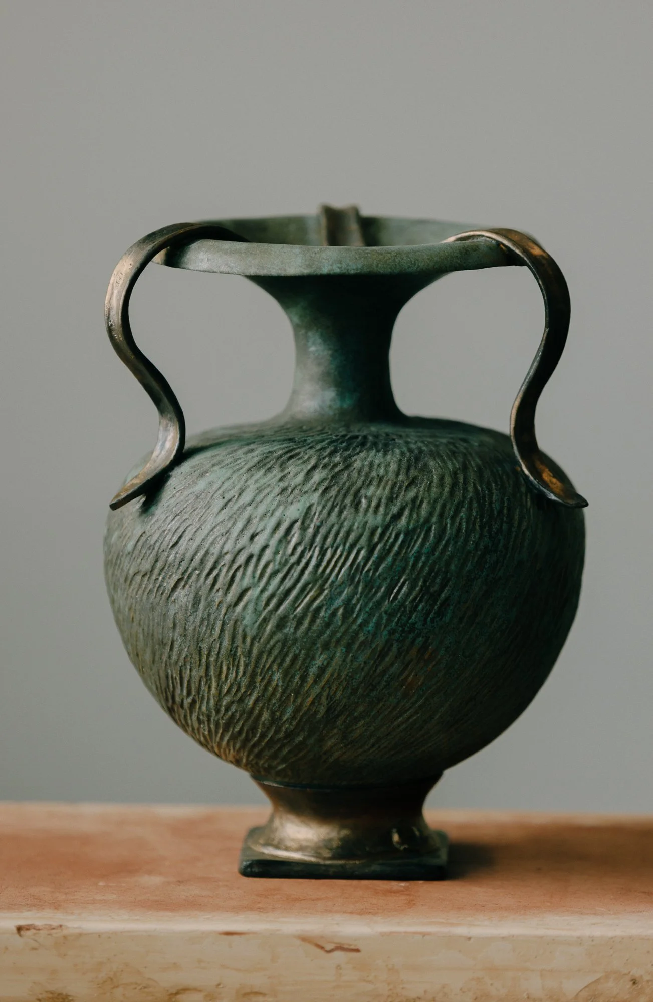 A decorative ceramic vase with a textured surface, a wide body, three curved handles, and a narrow neck, placed on a plaster surface.