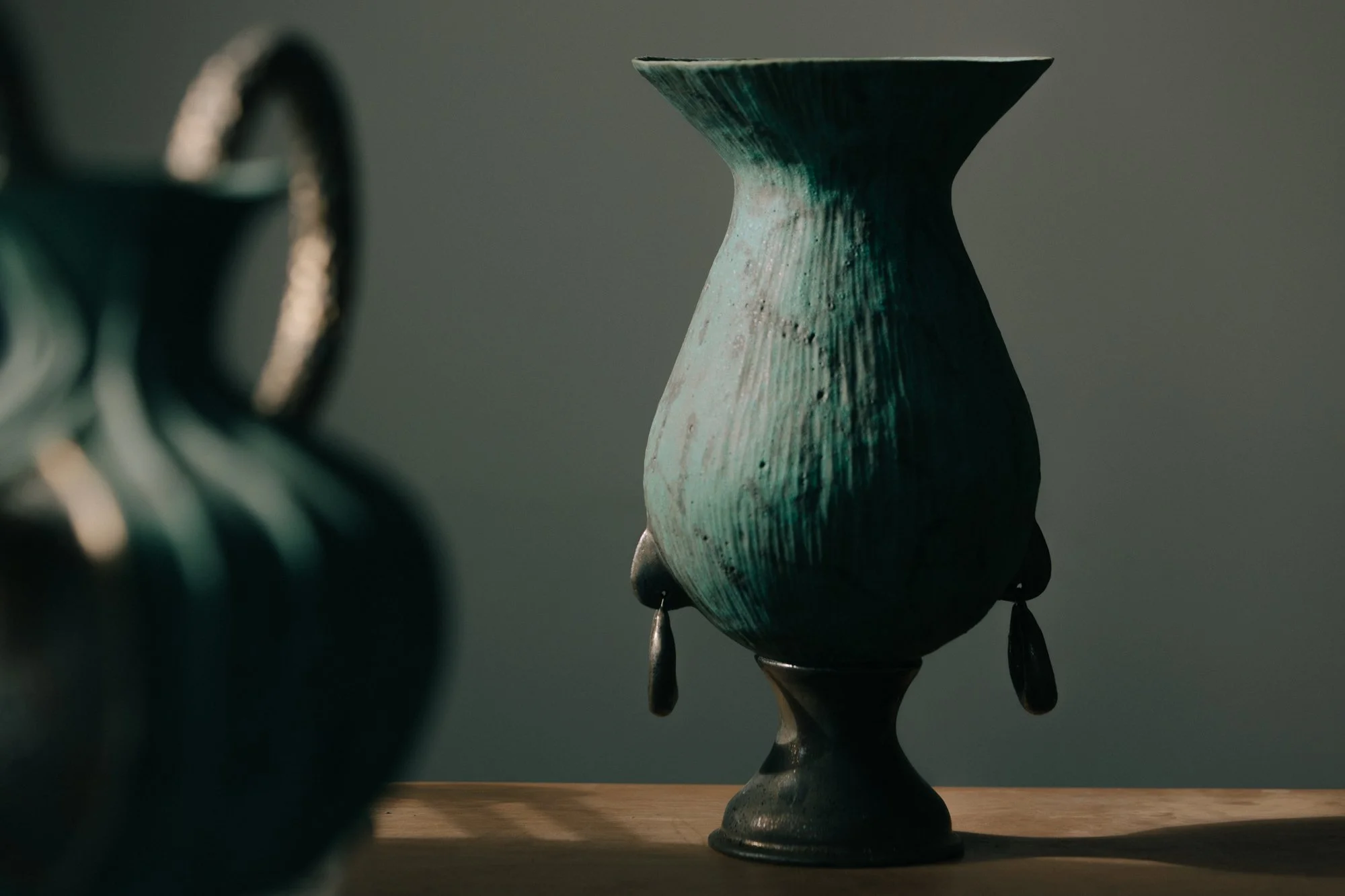 A close-up of a textured, greenish-blue decorative vase with dangling black ornaments, placed on a wooden surface with a plain wall background.