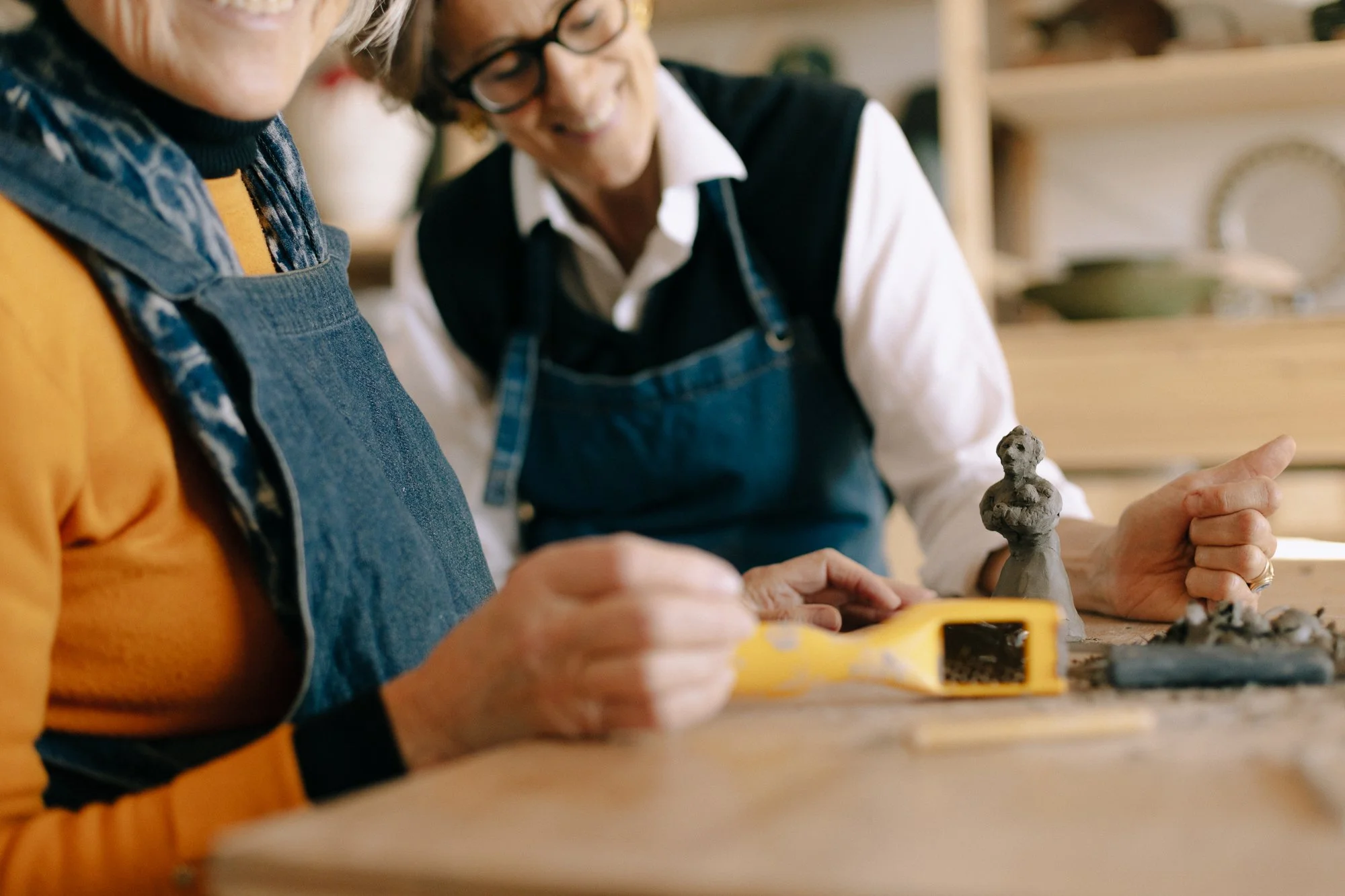Two women working with clay on a table, one using a yellow tool, with ceramic figures and pottery tools around them.