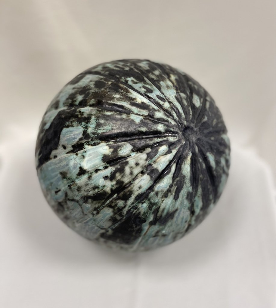 A close-up of a round, ceramic sculpture with a textured surface and a mix of dark and light marbled colors, placed against a white background.