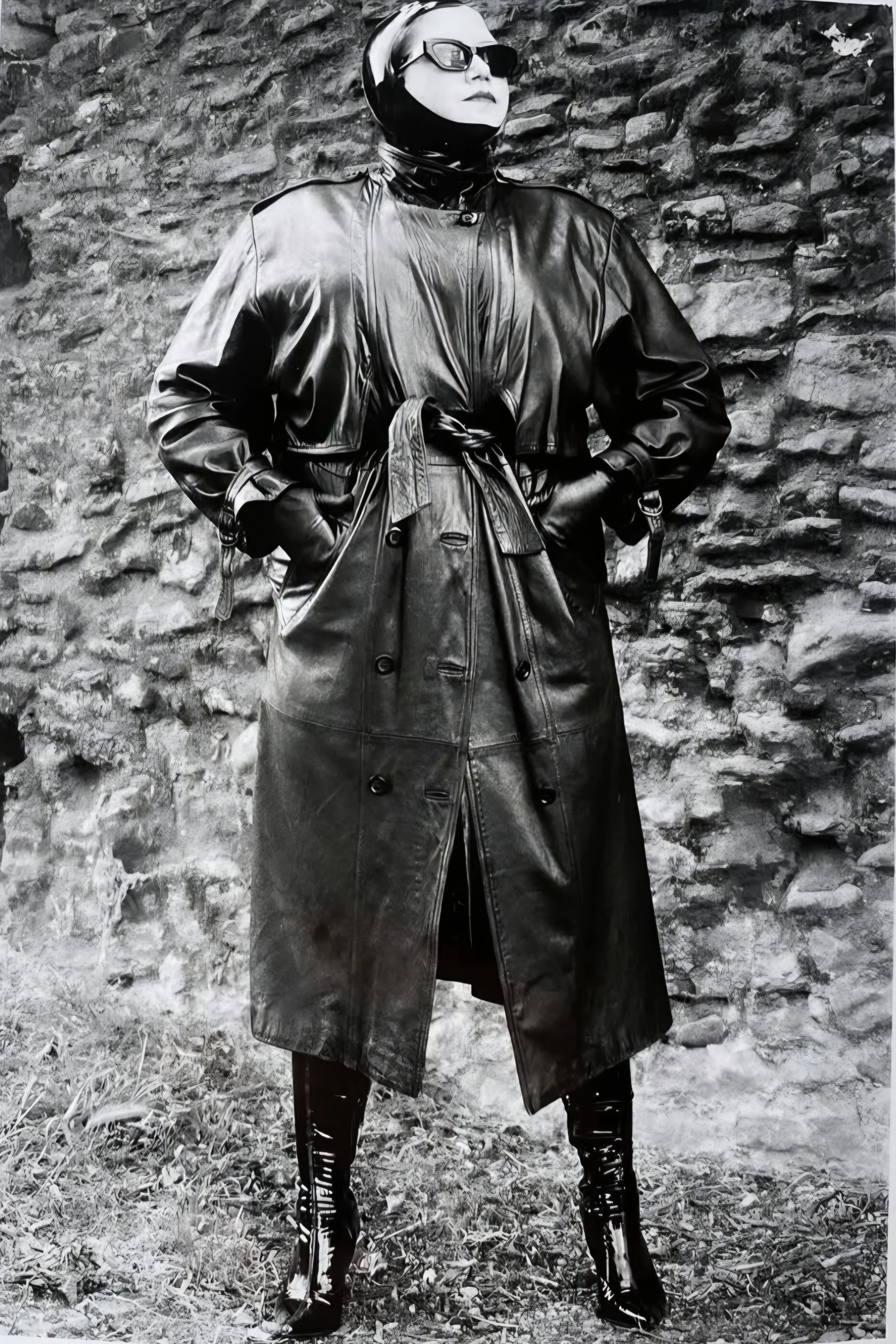 A female dominatrix in a long leather trench coat, wearing sunglasses and gloves, standing outdoors against a stone wall, with her hands in her coat pockets.