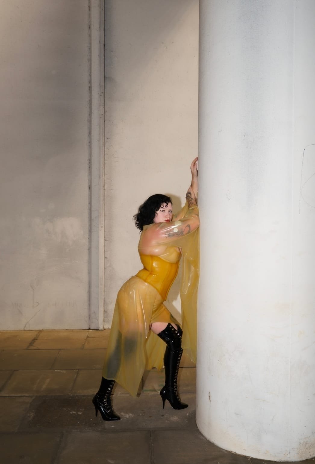 Dominatrix with black curly hair and tattoos wearing a sheer latex dress and high-heeled black boots, posing against a large white pillar with an industrial background.