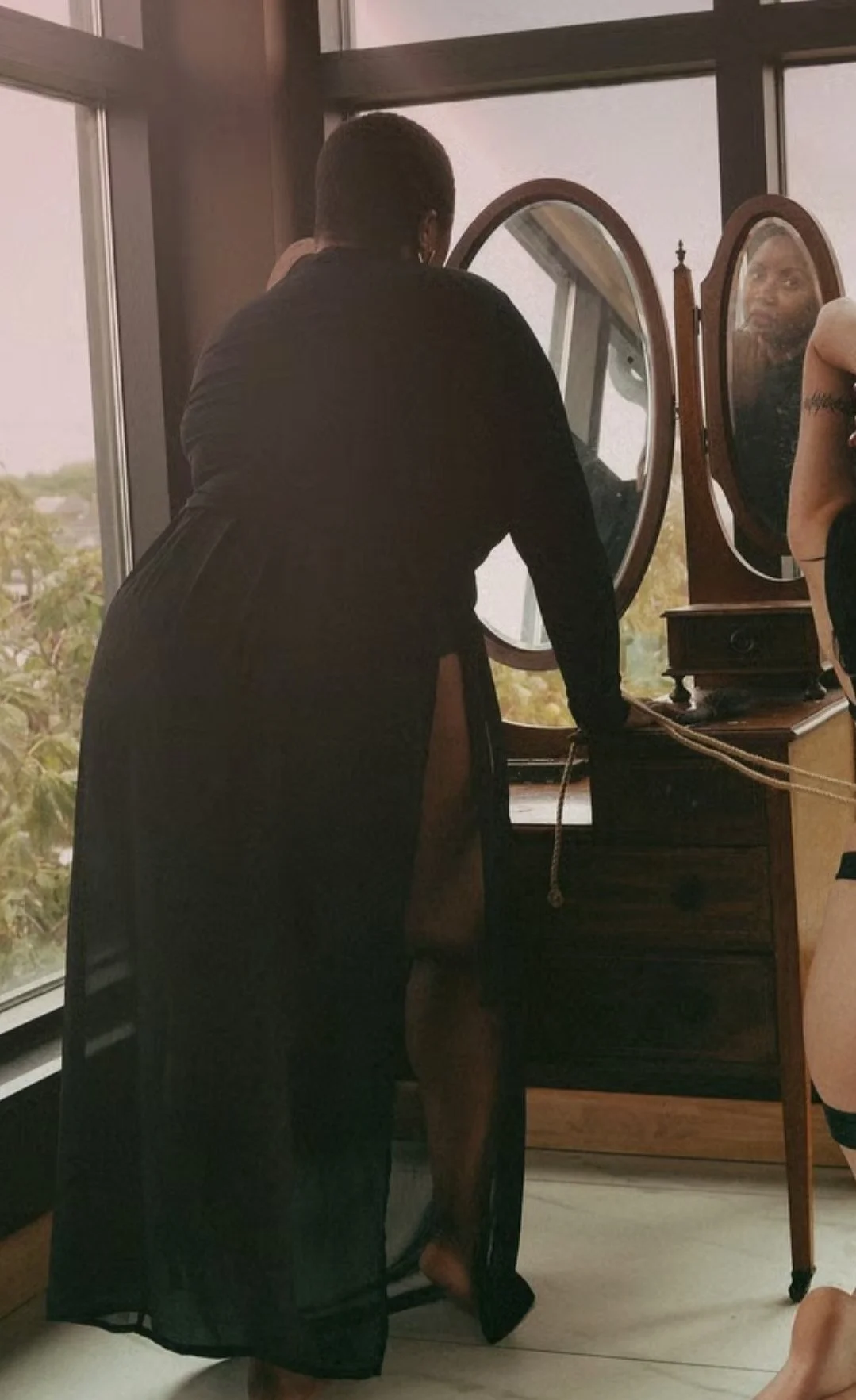A woman looking into a mirror on a wooden dresser in a room with large windows showing trees outside.