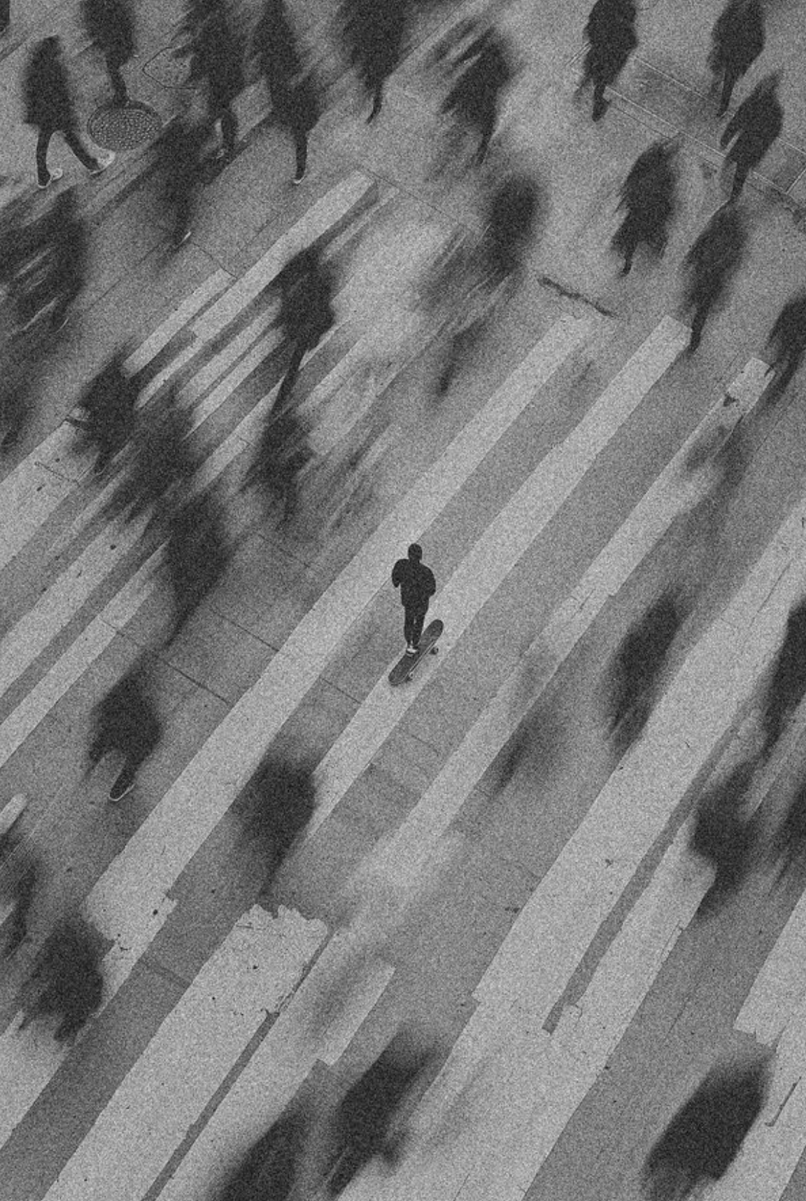 Black and white photo of a skateboarder on a crosswalk, surrounded by blurred figures walking in various directions.