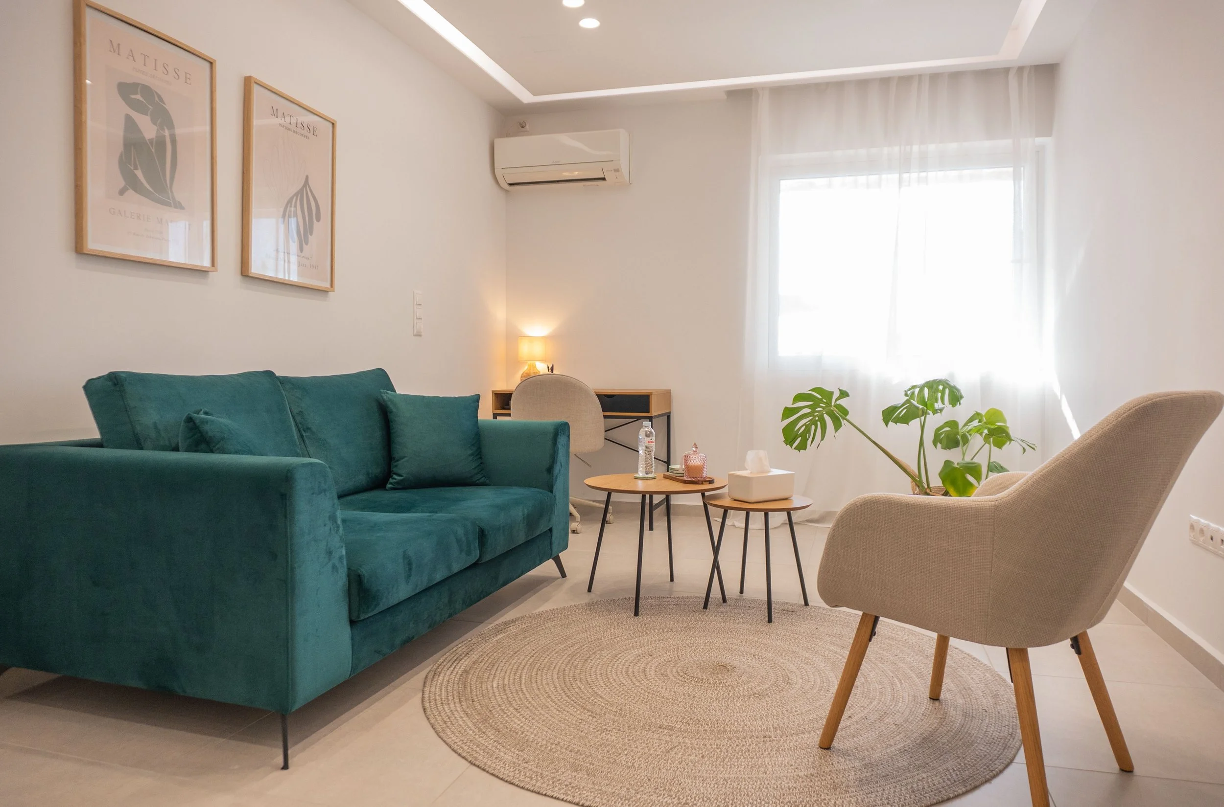 Living room with a teal sofa, beige armchair, small round tables, potted plant, wall art, window with white curtains, air conditioning unit, and a small desk with a lamp.