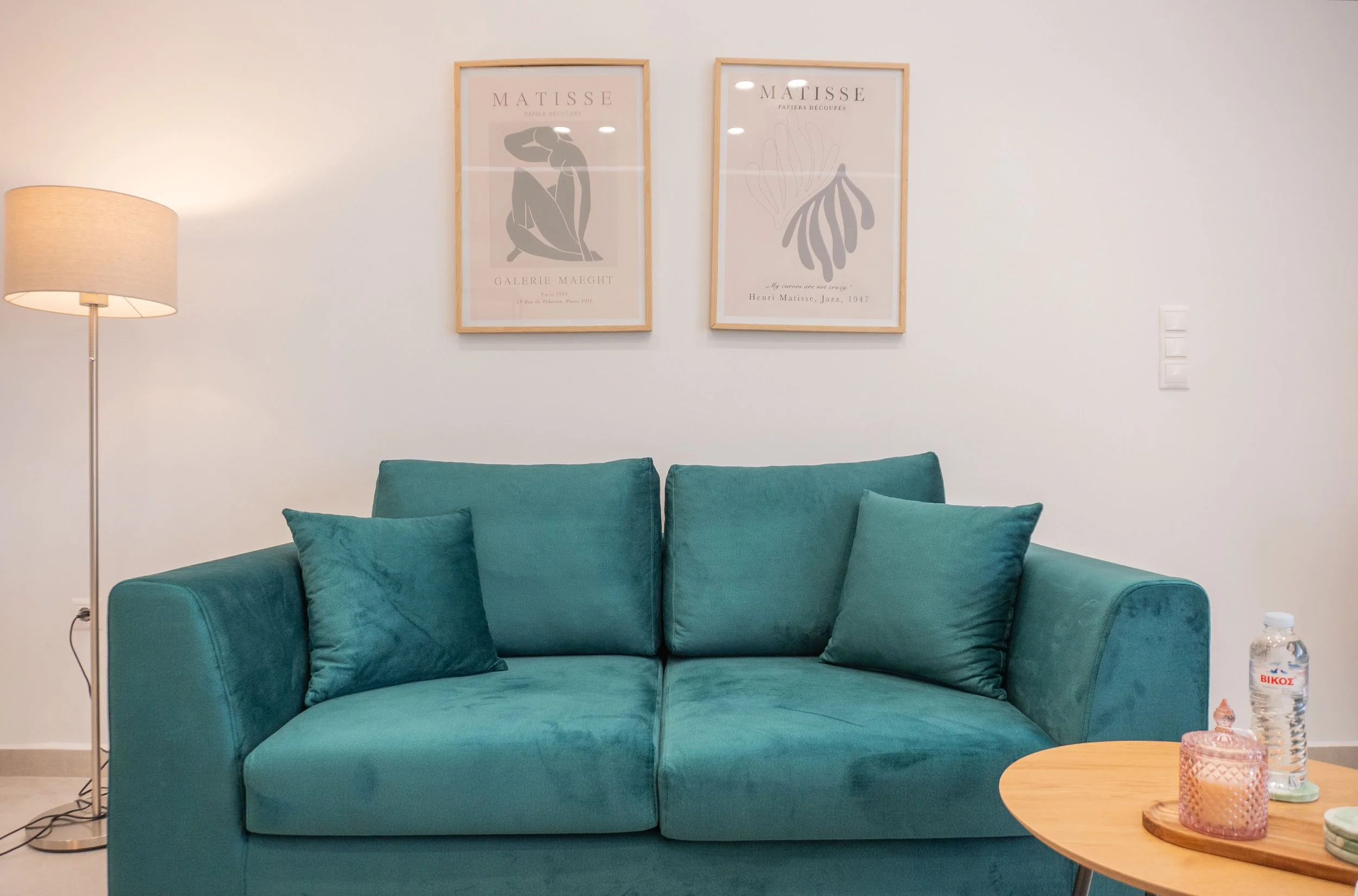 A teal sofa with two cushions in front of a white wall with two framed artworks, a floor lamp on the left, and a small wooden coffee table on the right holding a water bottle and candles.