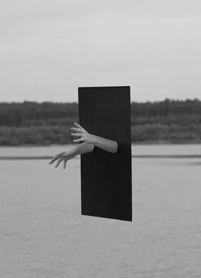 A person is behind a black rectangular panel, with only their hands and part of their arm visible, sticking out as if reaching through the panel in a surreal outdoor setting.