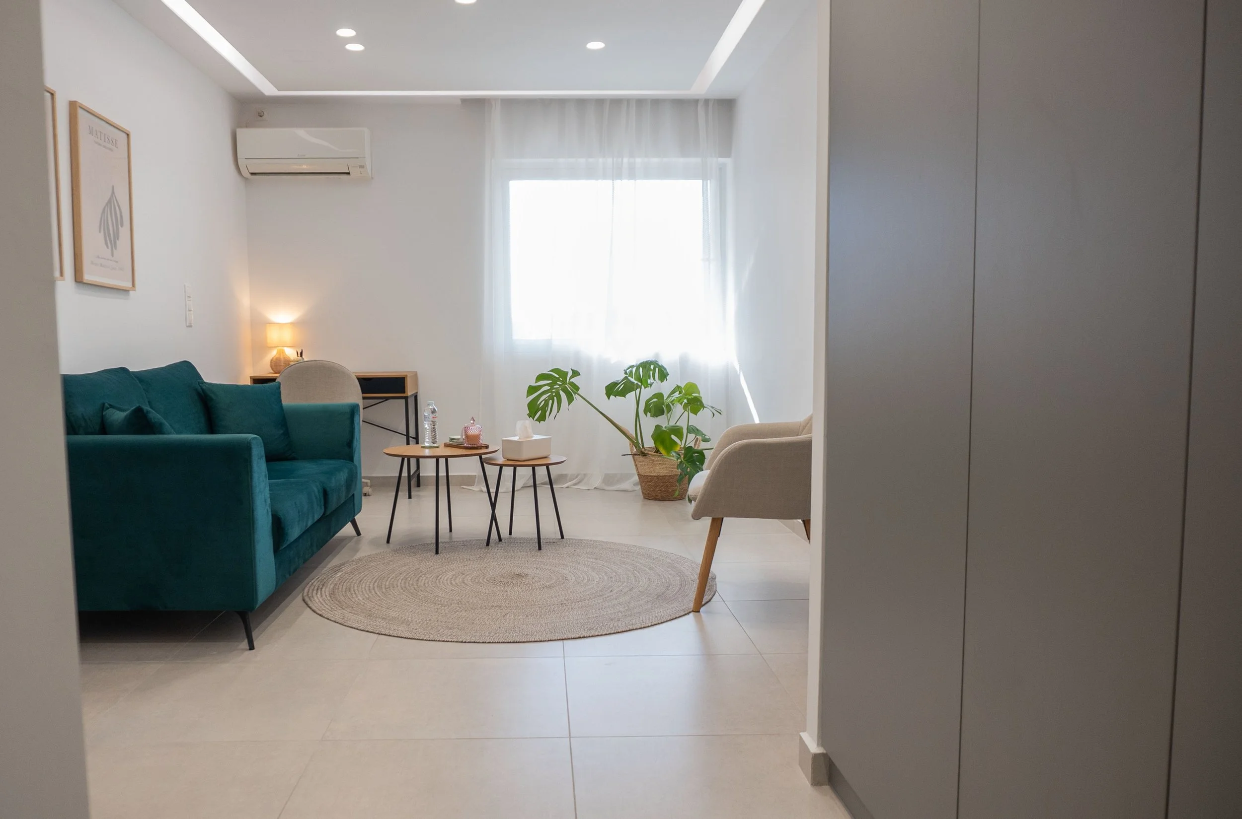 Living room with a teal sofa, beige armchair, small round tables, a window with curtain, a potted plant, a rug, wall art, and an air conditioning unit.