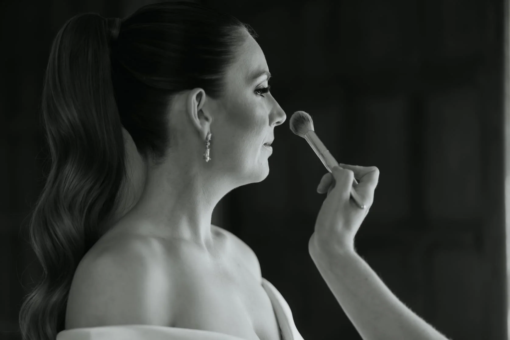 A woman with wavy hair and earrings is applying makeup with a brush in front of a mirror.
