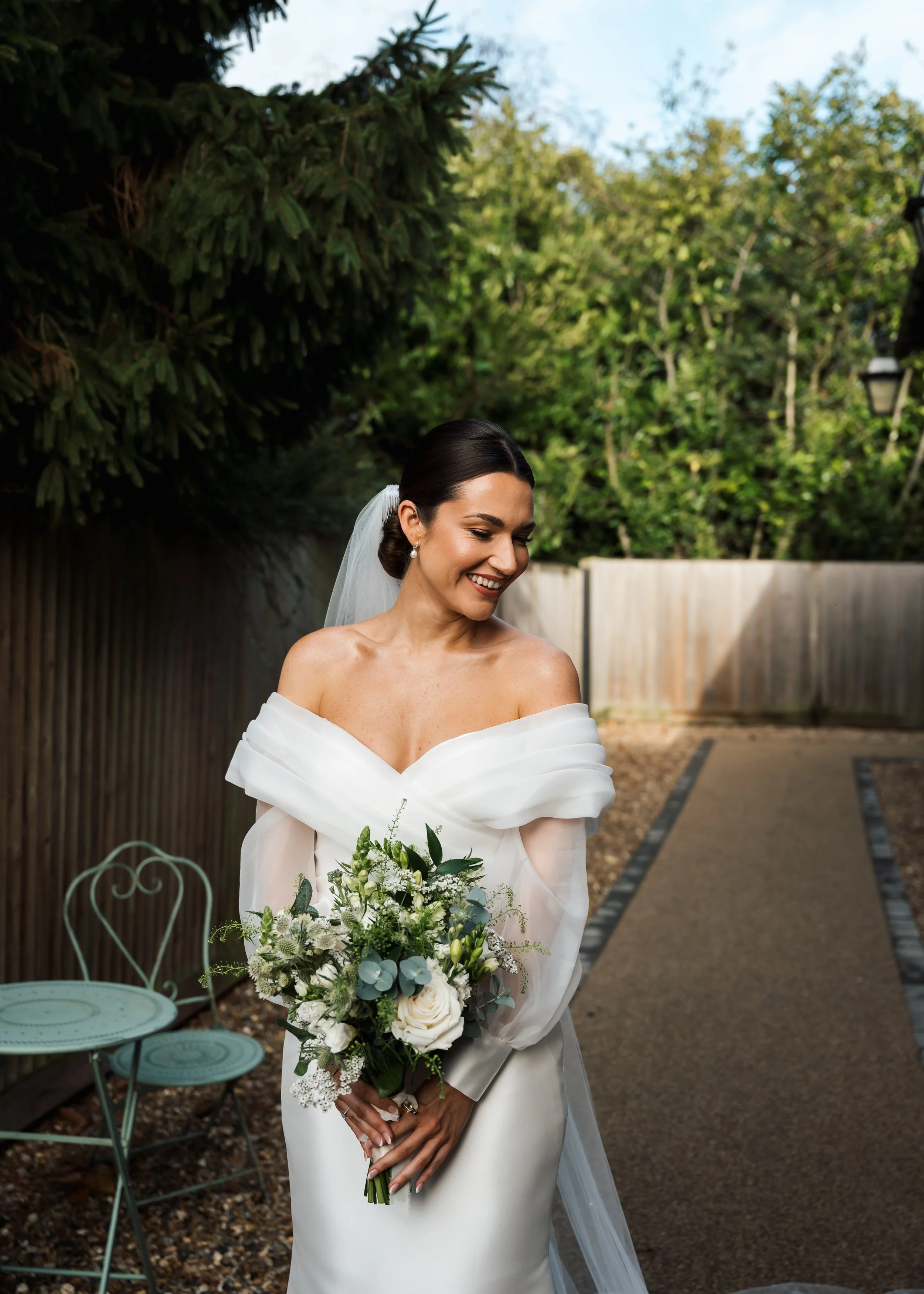 A bride in a white off-the-shoulder wedding dress holding a bouquet of white flowers and greenery, smiling and looking down in an outdoor garden setting with trees, a wooden fence, and vintage-style garden furniture.