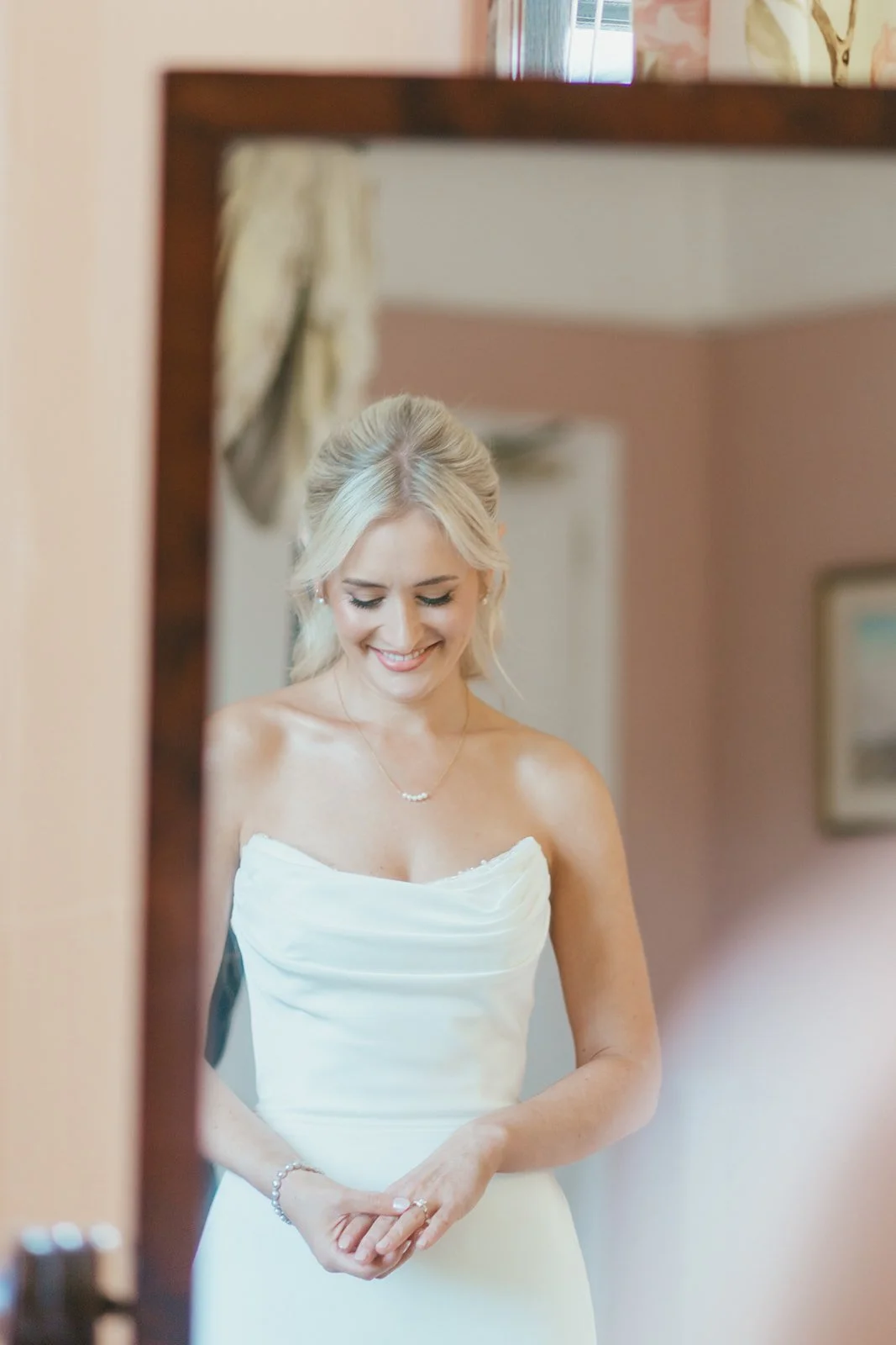 A smiling blonde bride in a white strapless wedding dress, standing indoors, reflected in a mirror.