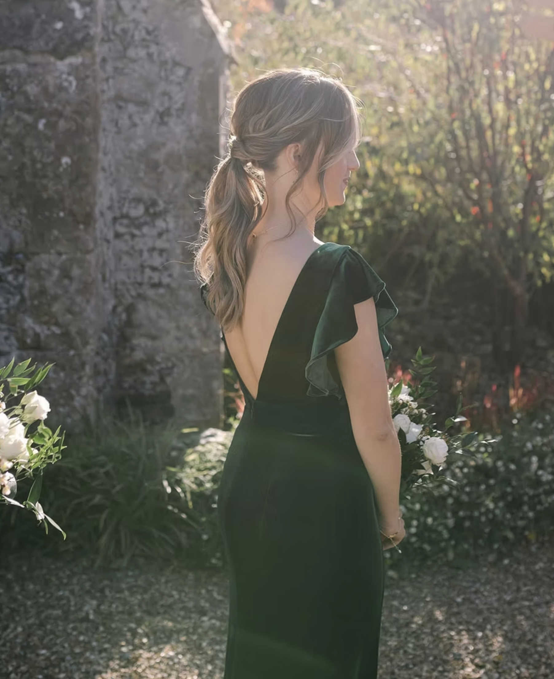 A woman with long wavy hair in a loose braid, wearing a dark dress with a V-shaped back, standing outdoors near a stone wall, surrounded by greenery and flowers, with sunlight creating a soft glow.