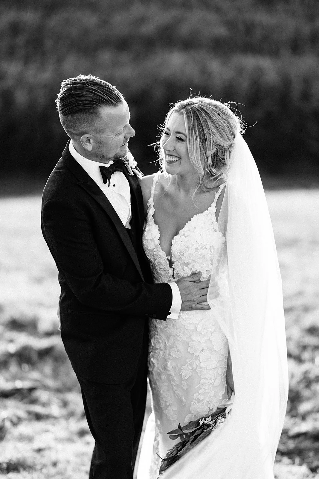 A bride and groom smiling and looking at each other outdoors during their wedding, with the groom in a tuxedo and the bride in a lace wedding dress holding a bouquet.