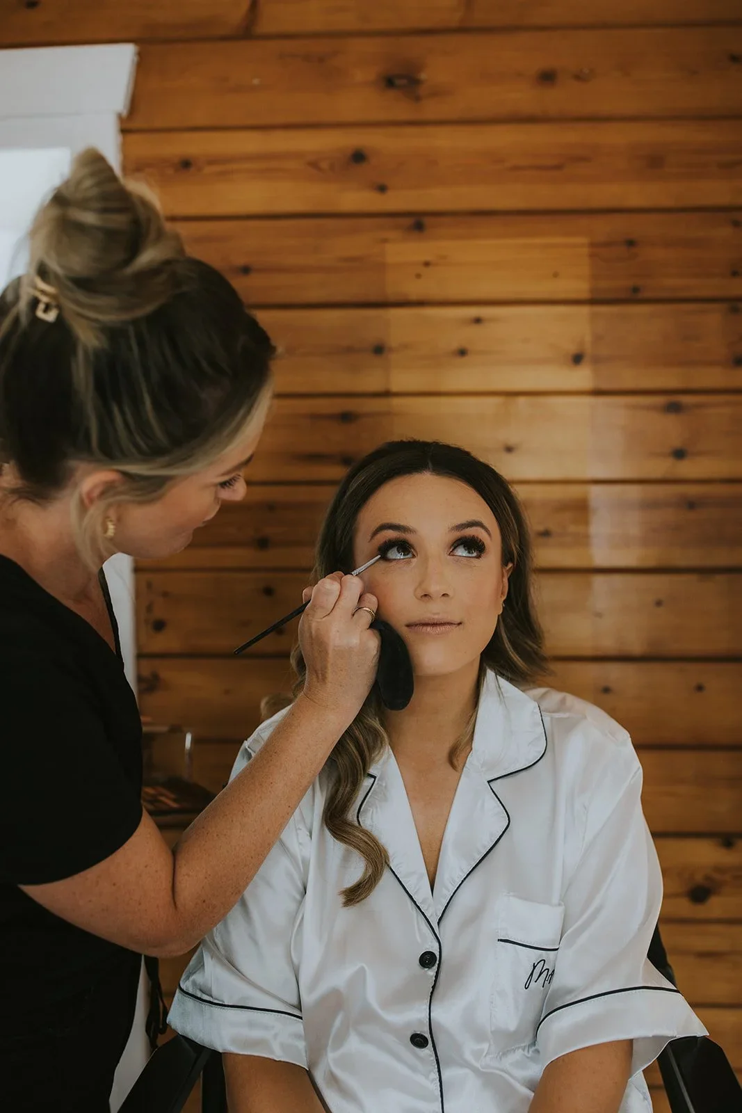 A woman is sitting in a chair with pajamas, having makeup applied by a makeup artist. The makeup artist is applying eyeshadow or eyeliner to the woman's eye. The setting has wooden walls in the background.