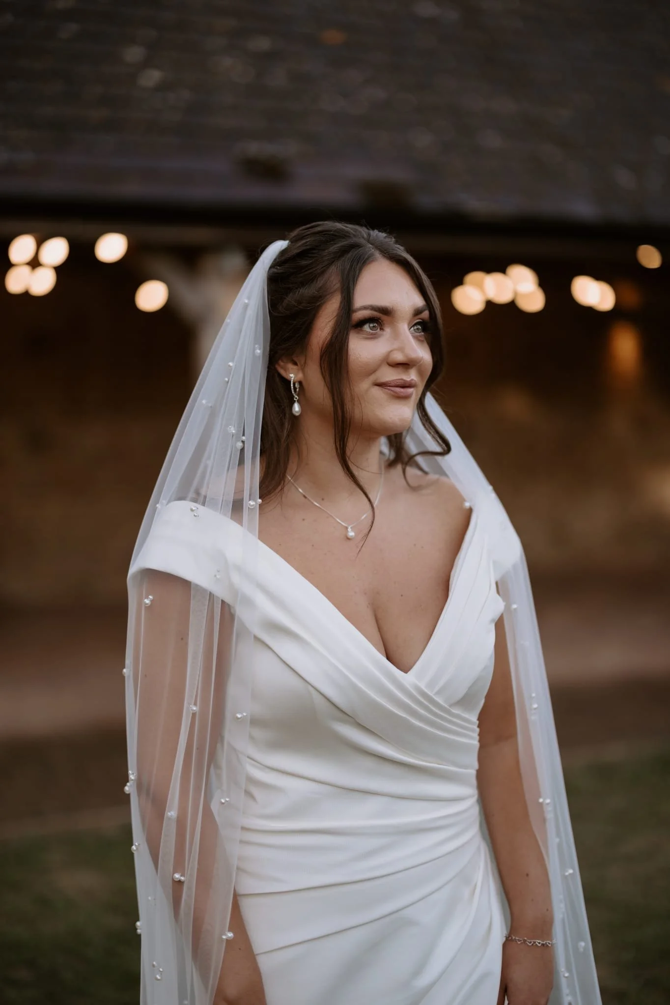 A woman in a white wedding dress with a veil and jewelry stands outdoors, with blurred background lighting.