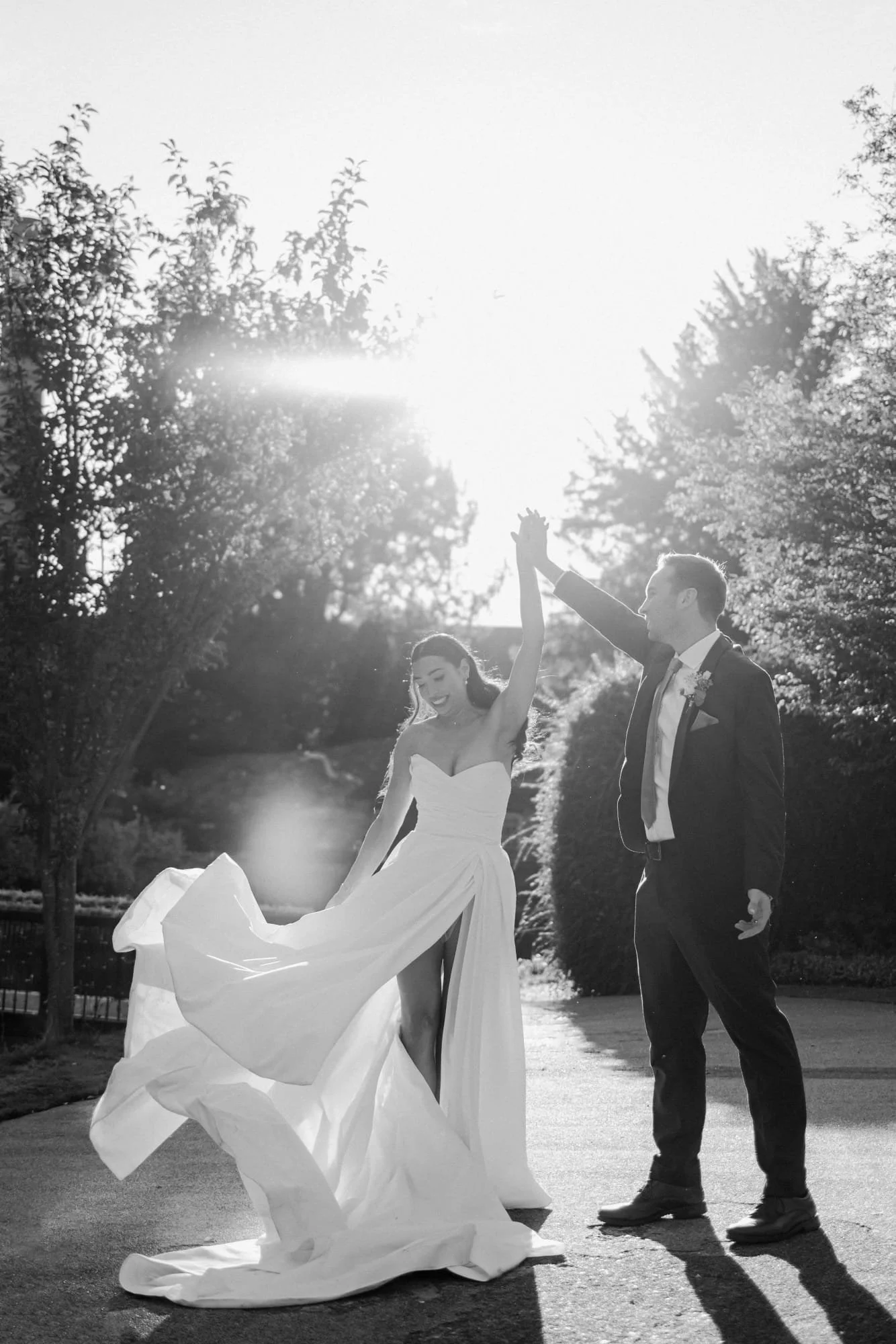 A bride and groom hold hands and dance outdoors on a sunny day, with the bride in a strapless wedding gown and the groom in a dark suit, surrounded by trees.