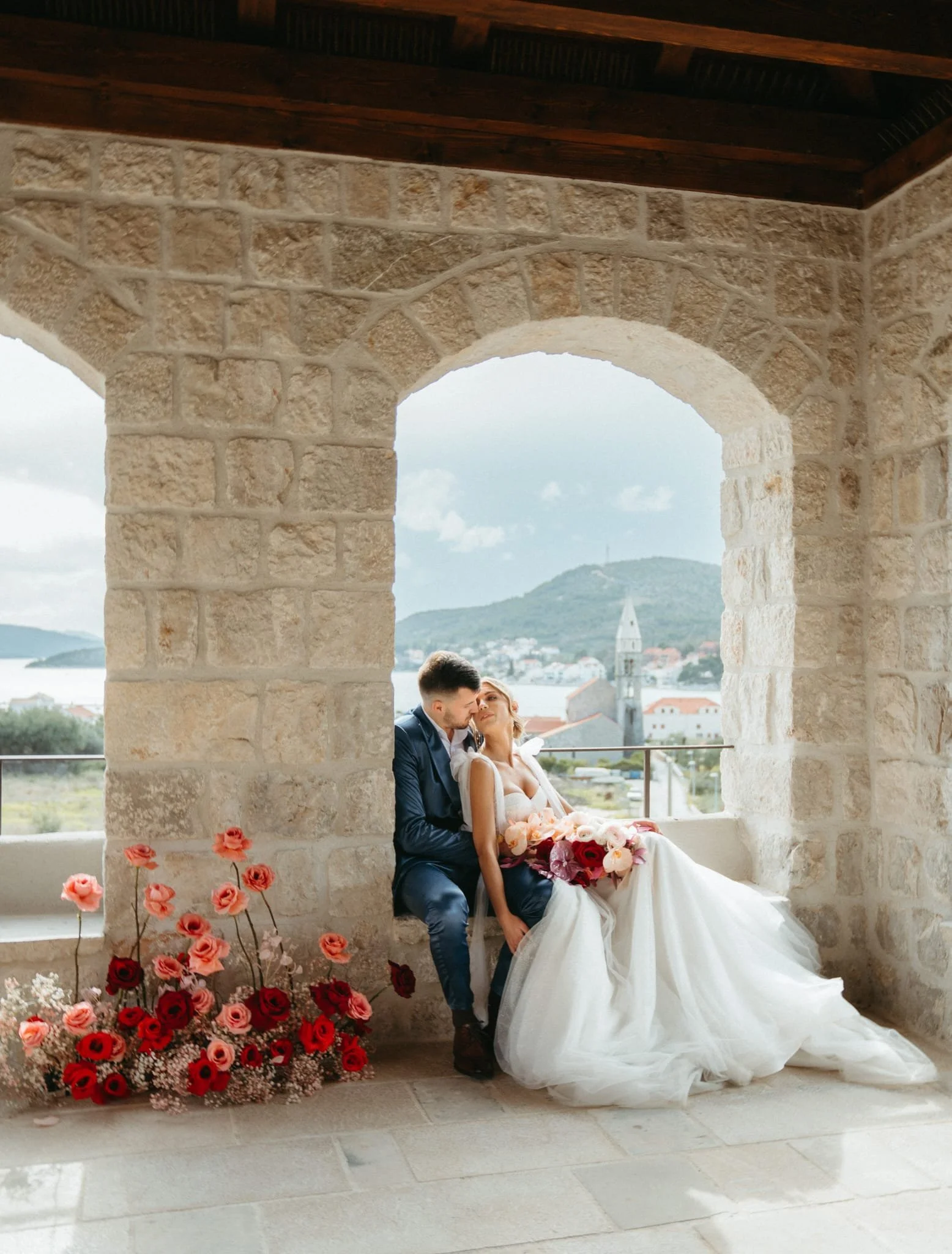 A newly married couple sits under a stone archway with a view of a town and mountains in the background. The woman wears a white wedding dress and holds a bouquet of pink and red flowers. The man wears a dark suit and is leaning towards the woman. Pink and red flowers are arranged at the base of the arch.