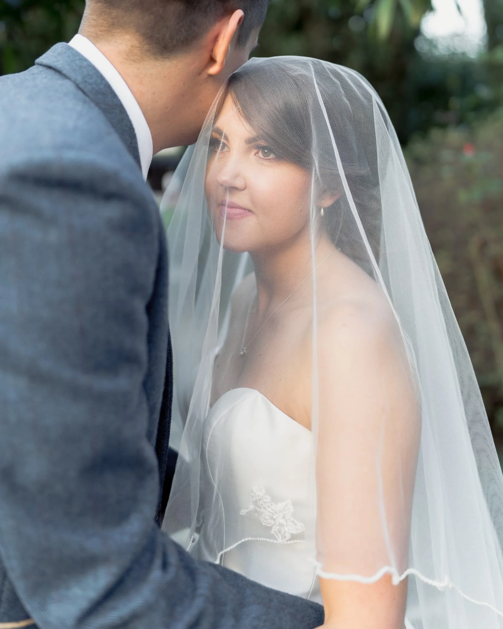 A bride and groom sharing a kiss at their wedding, with the bride wearing a veil and white dress, and the groom in a gray suit, outdoors with blurred greenery in the background.