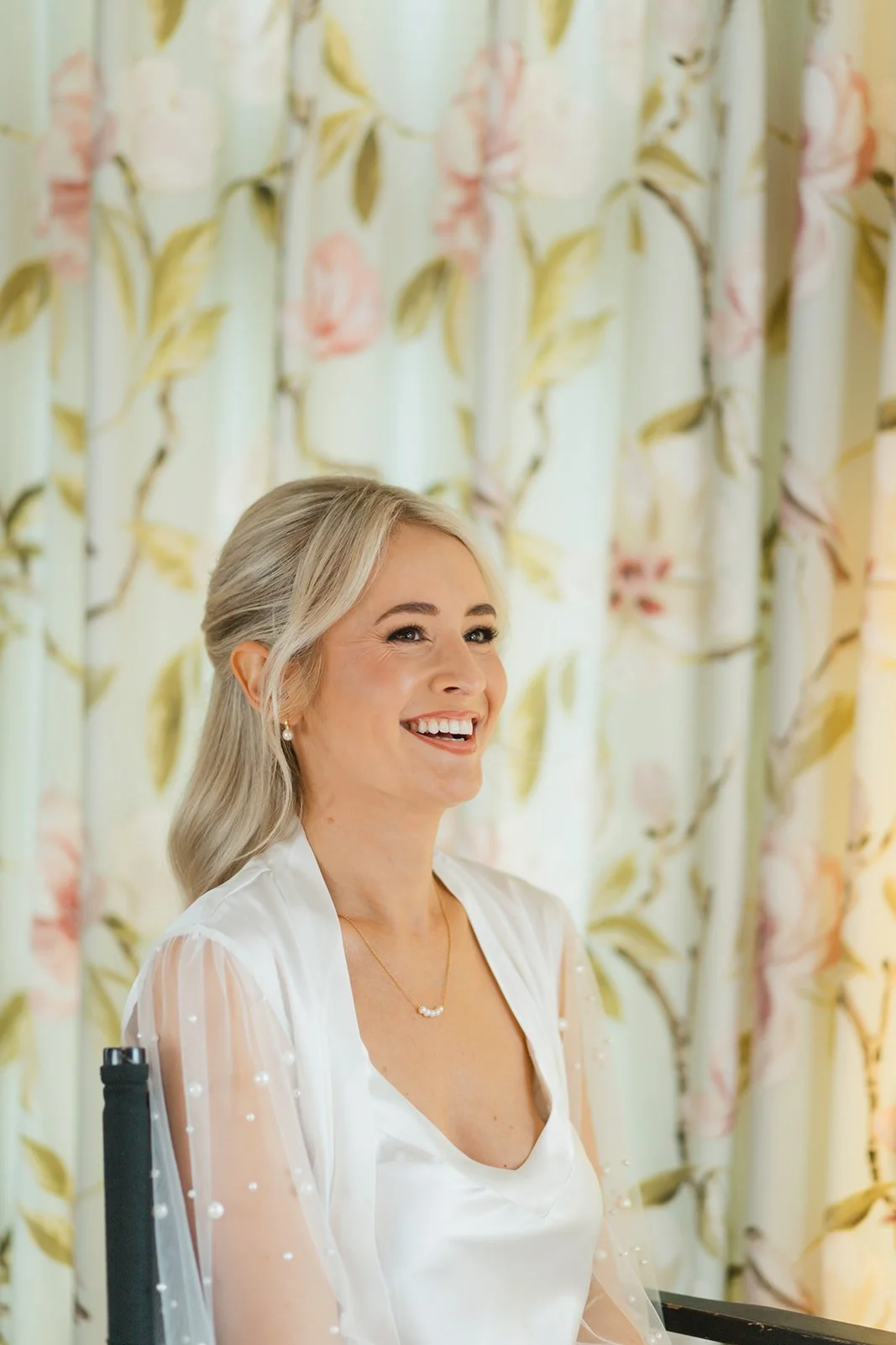 A middle-aged woman with blonde hair styled in soft waves, smiling joyfully, wearing a white satin top with sheer puffed sleeves adorned with tiny pearls, a delicate necklace, and pearl earrings, seated in front of floral curtains with pink and yello