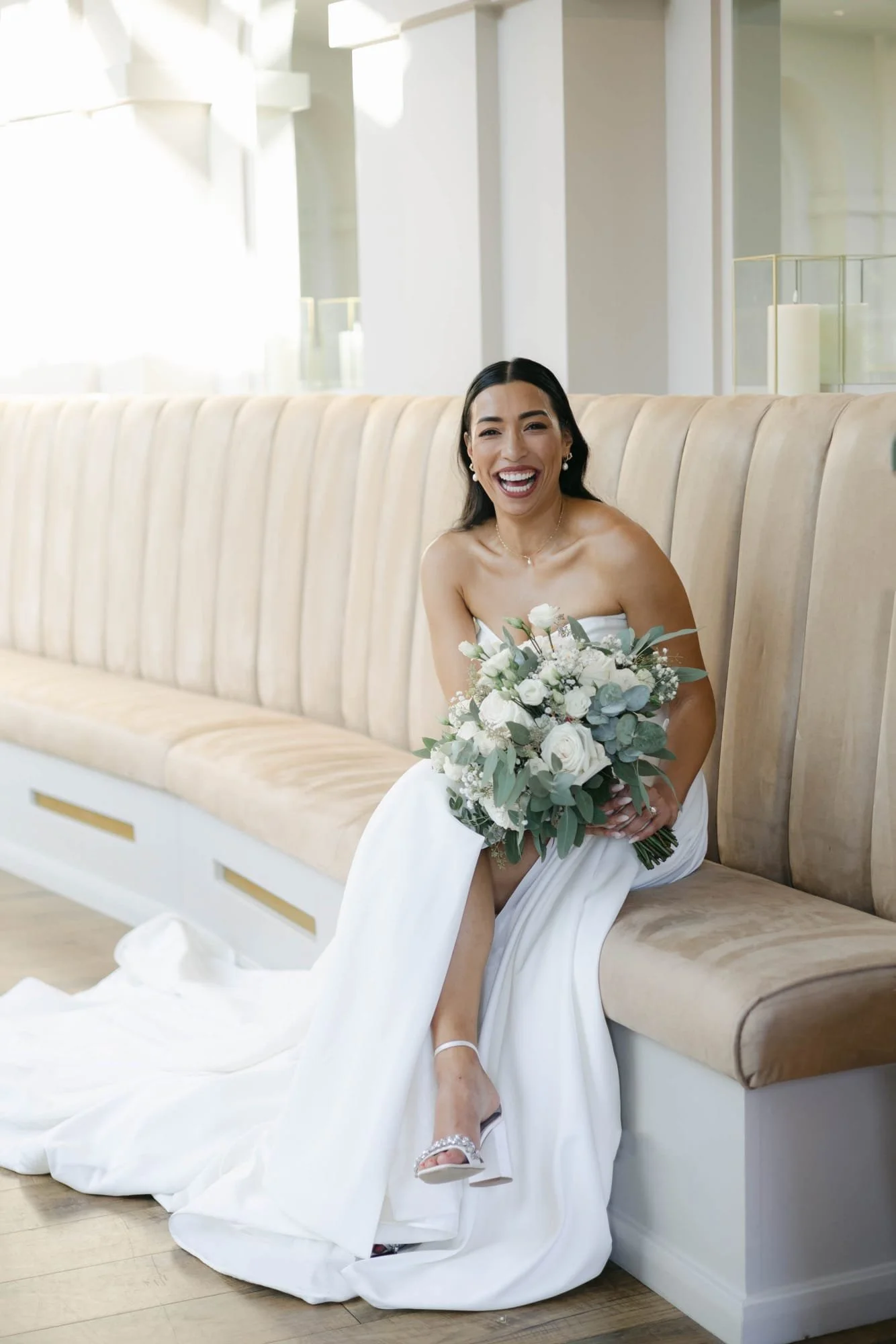 A woman in a wedding dress sitting on a beige velvet bench, holding a bouquet of white flowers and greenery, smiling and laughing.