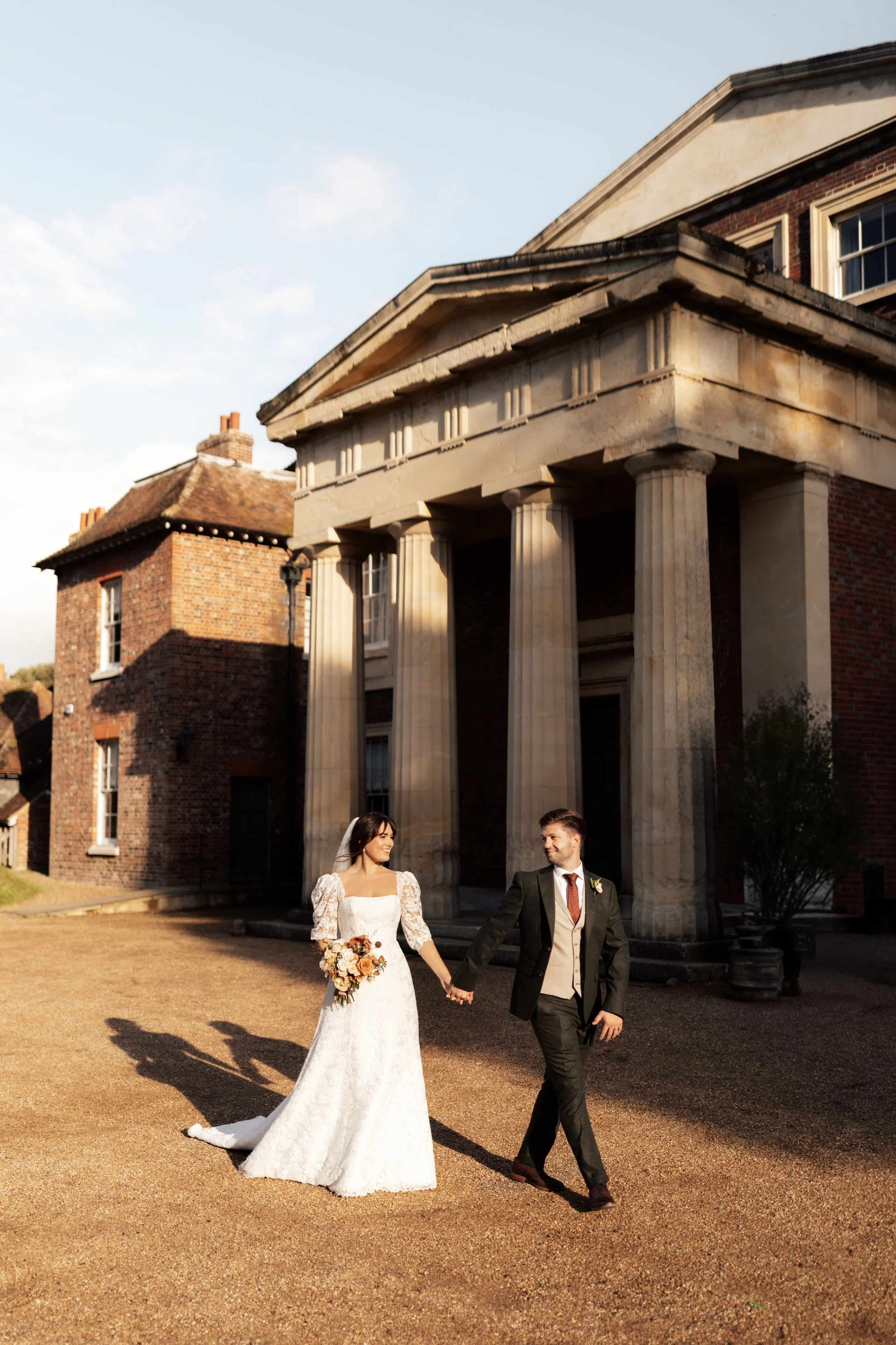 A bride and groom walking hand in hand outside a historic building with columns, during their wedding day.