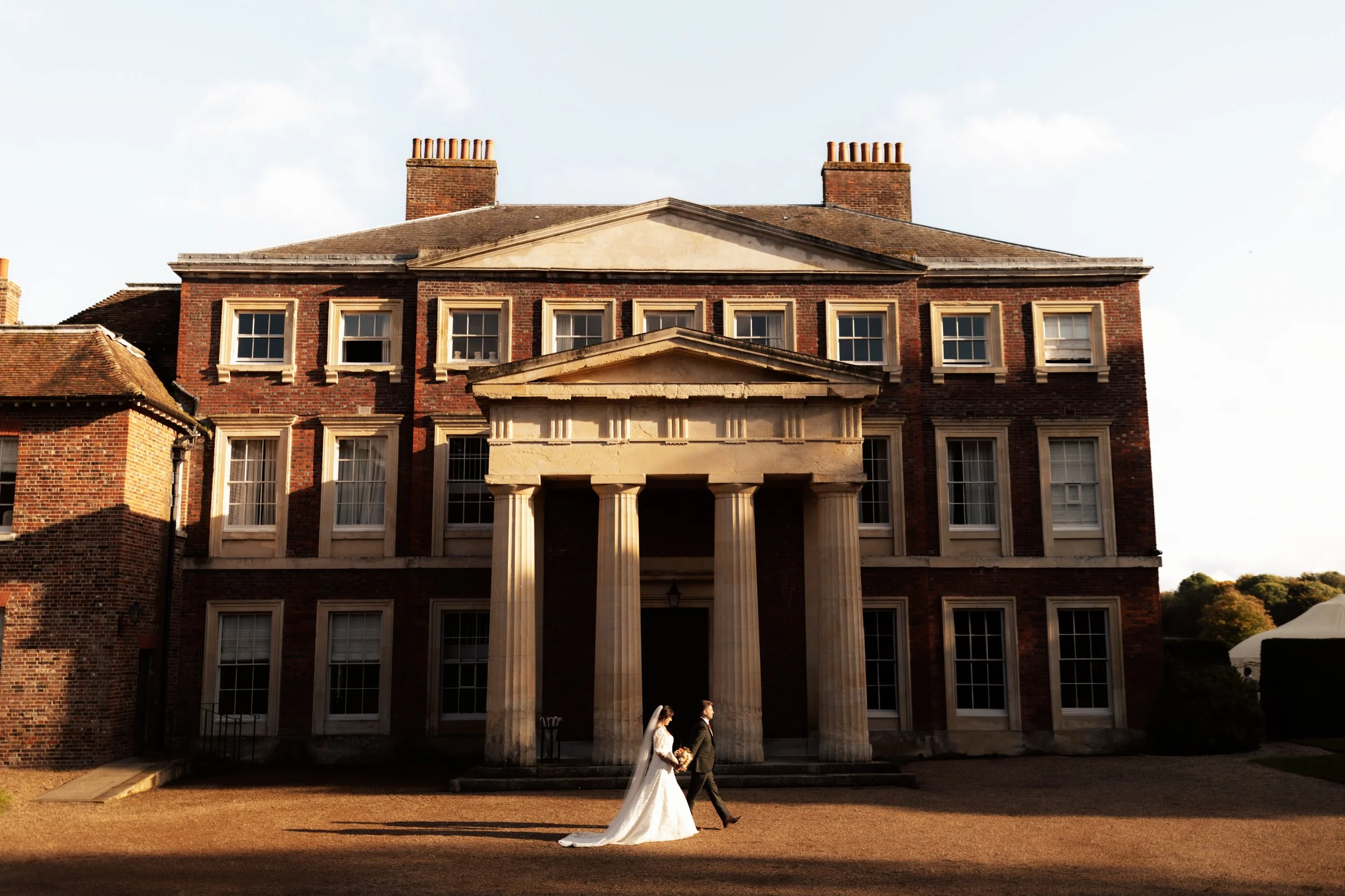 Bride and groom walking in front of a large brick mansion with columns, under a cloudy sky during daylight.