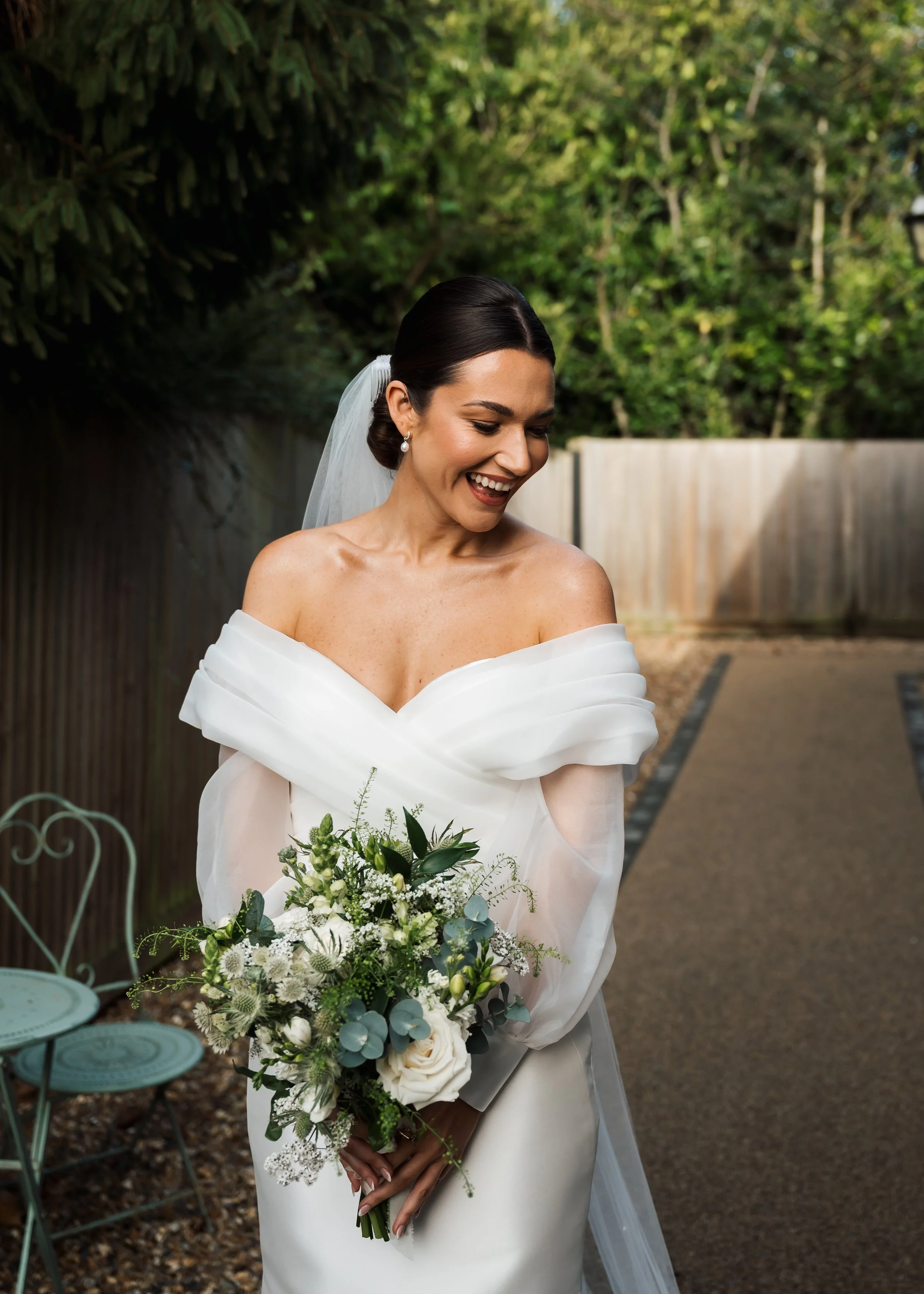 A bride in an off-the-shoulder white wedding dress holding a bouquet of white and green flowers, smiling outdoors with green trees and a wooden fence in the background.
