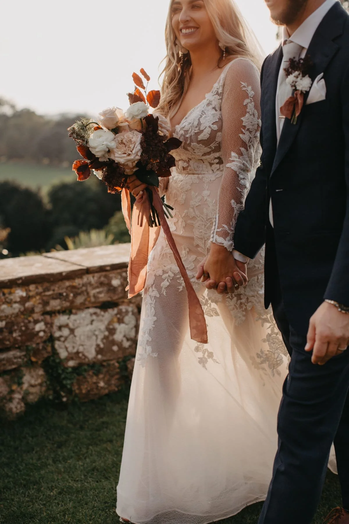 A bride and groom holding hands during their wedding ceremony outdoors, with the bride wearing a lace gown and holding a bouquet of roses and foliage, at sunset.