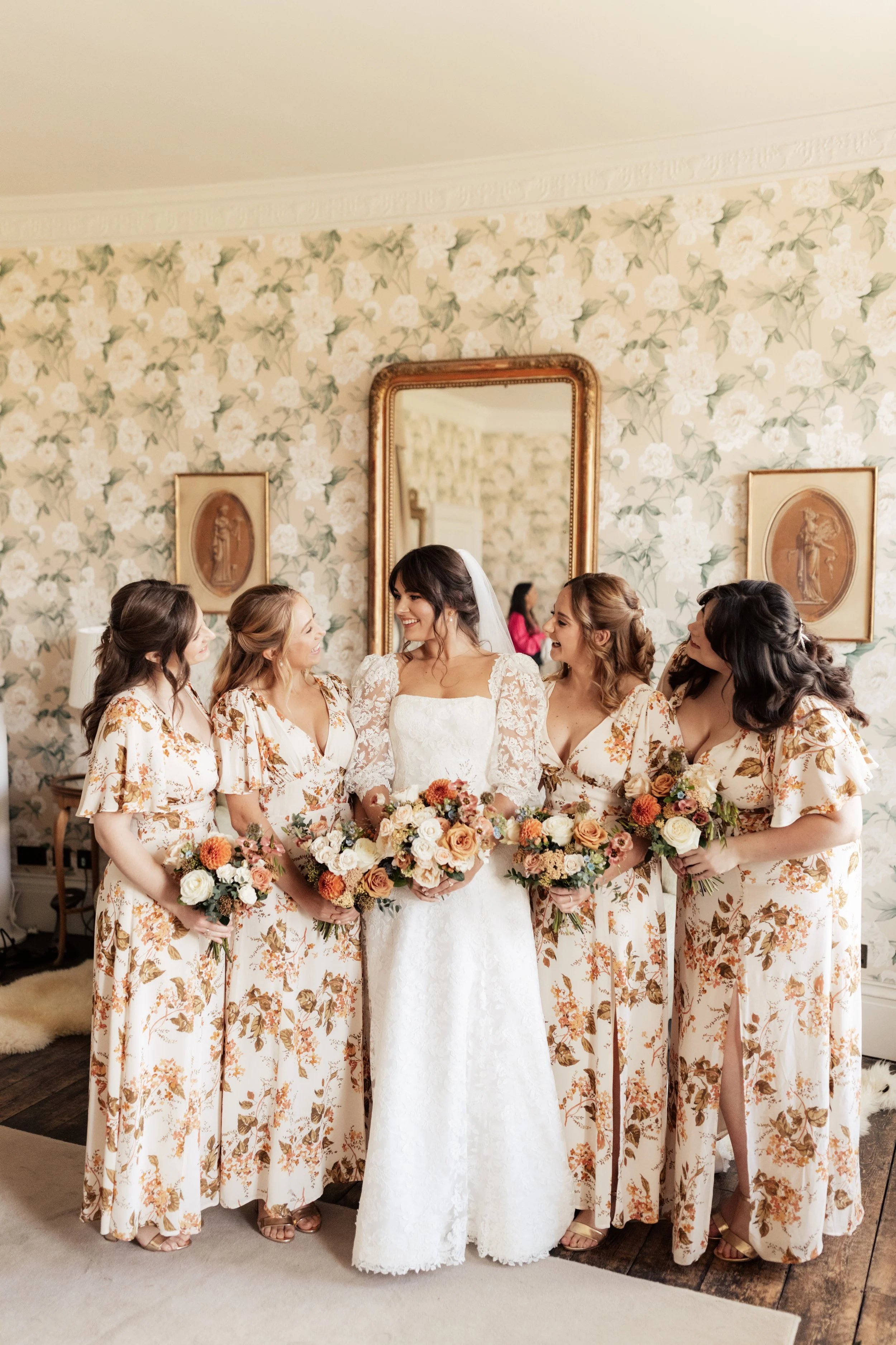 Bridal party, including the bride and five bridesmaids, all holding bouquets, standing indoors in front of a floral wallpapered wall with a large mirror and framed artwork.