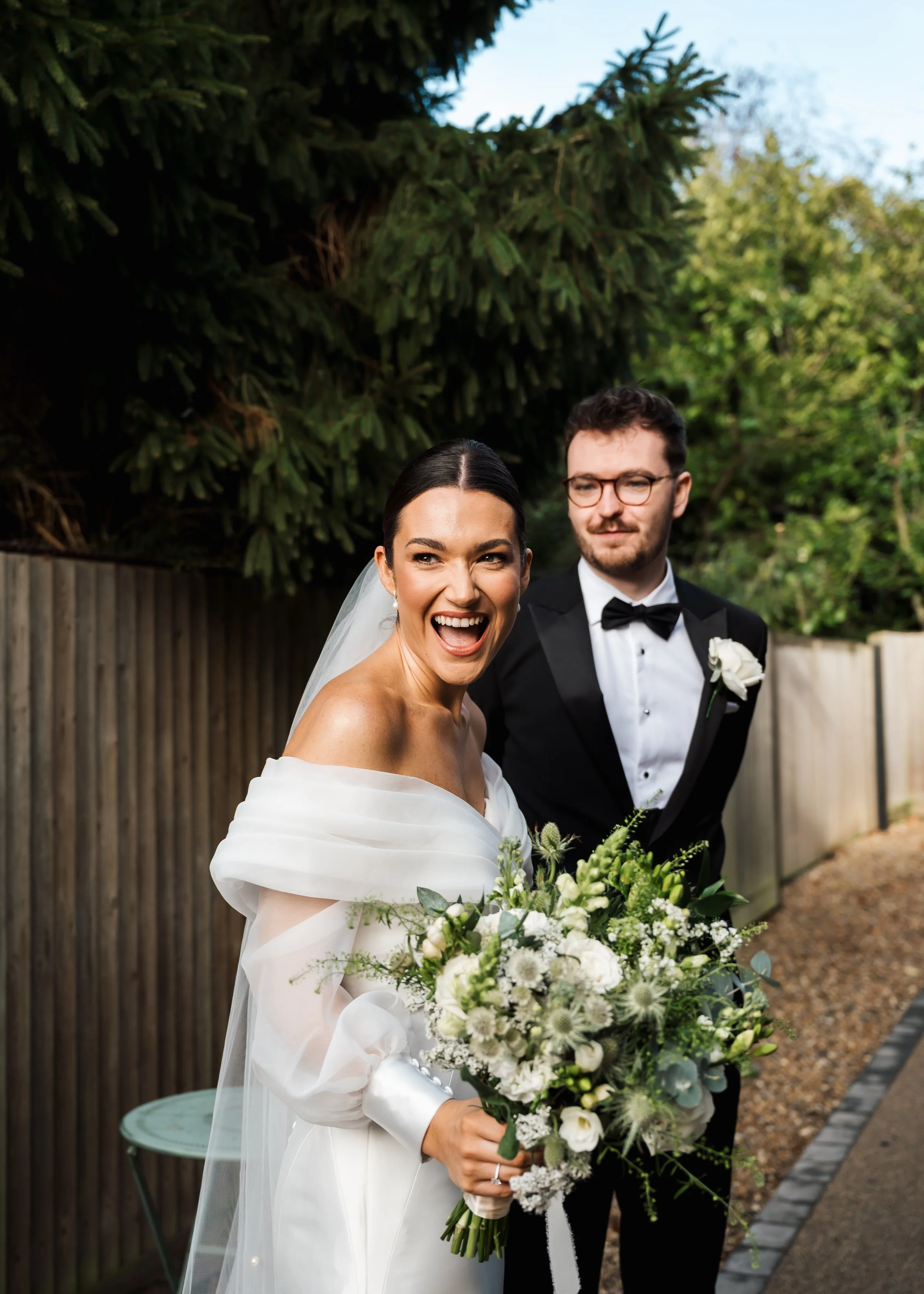 A bride holding a bouquet of white and green flowers, smiling excitedly, standing next to a groom in a black tuxedo outdoors with trees and a wooden fence in the background.